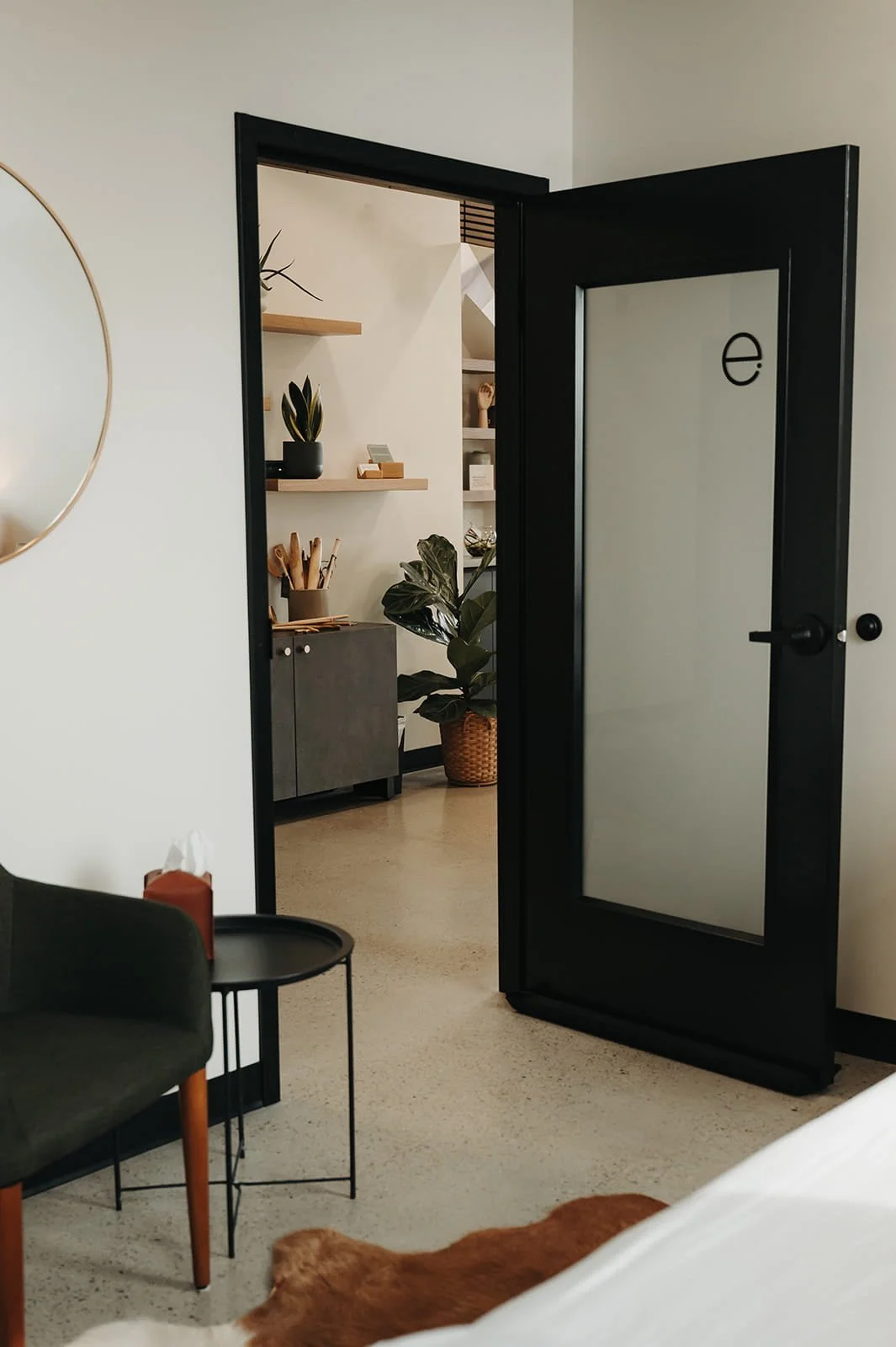 Interior view through a black-framed glass door showing a room with plants, shelves, and a black cabinet.