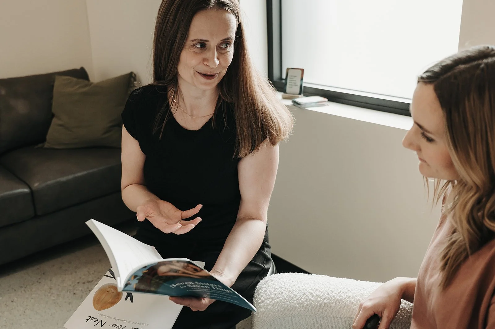 Two women having a conversation in a living room, one is holding a book and the other is seated with a blanket on her lap, with a window in the background.