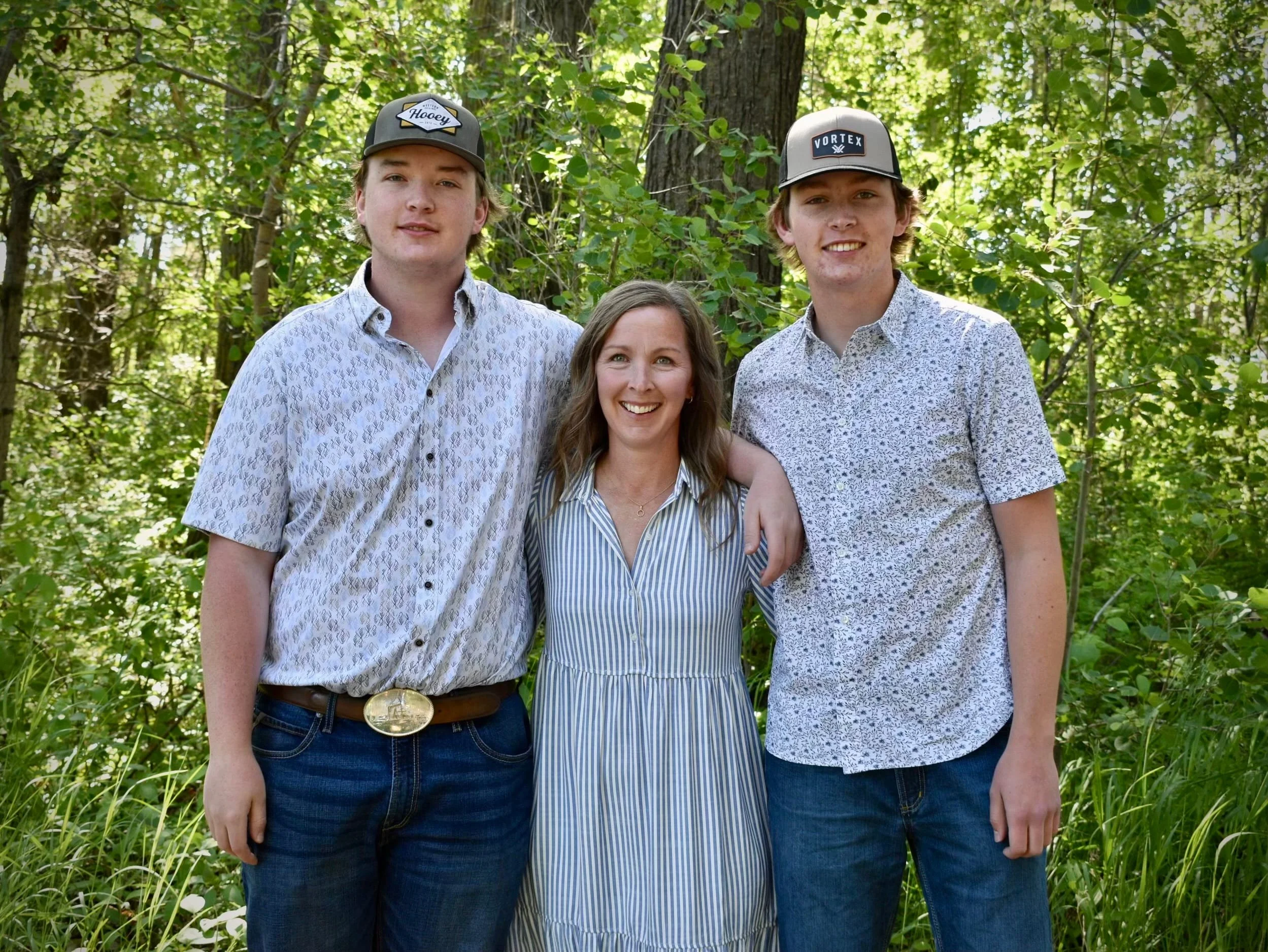 Three people, two young men and a woman, standing outdoors in a wooded area with green leaves, smiling at the camera.