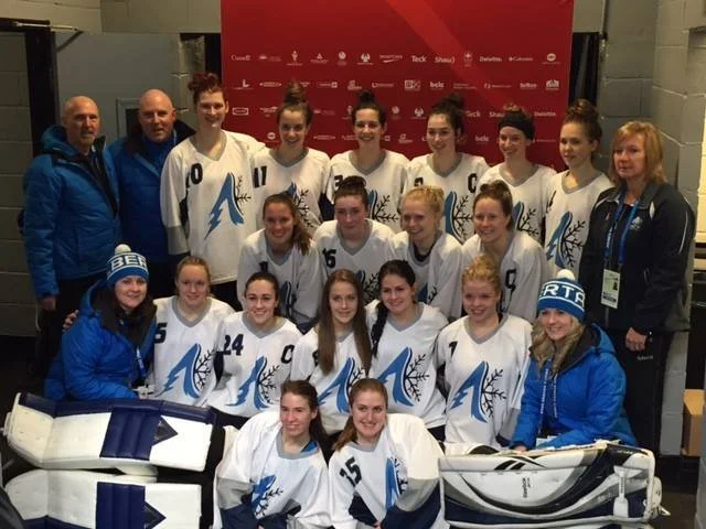 A women's ringette team in white jerseys with a blue logo, posing with their coaches and staff in front of a red backdrop with logos.