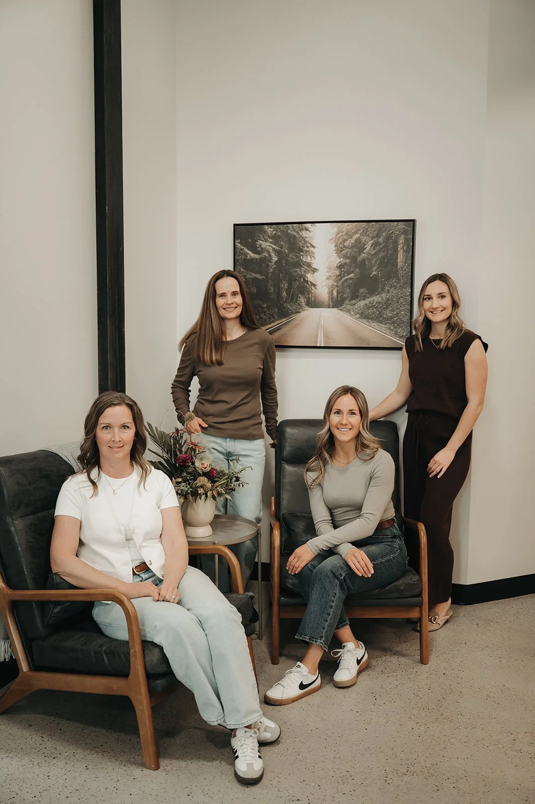 Four women in a room with a landscape photo of a road through a forest on the wall; one seated and three standing, all practitioners at Holm Collective, Lacombe AB.