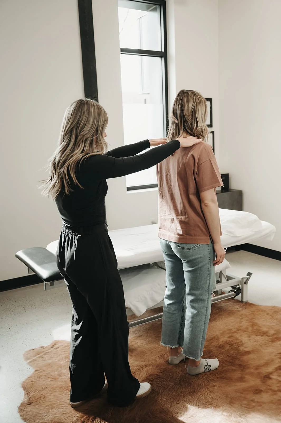 A woman in a brown t-shirt and jeans standing upright on a brown rug in a room with a medical examination bed, as a second woman in black clothing conducts an alignment or assessment.