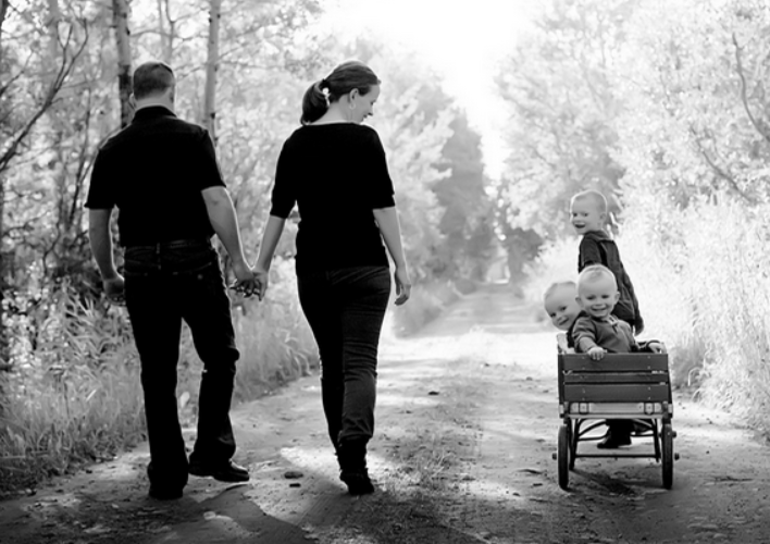 A family of four walking outdoors on a dirt path, with two children sitting in a wagon.