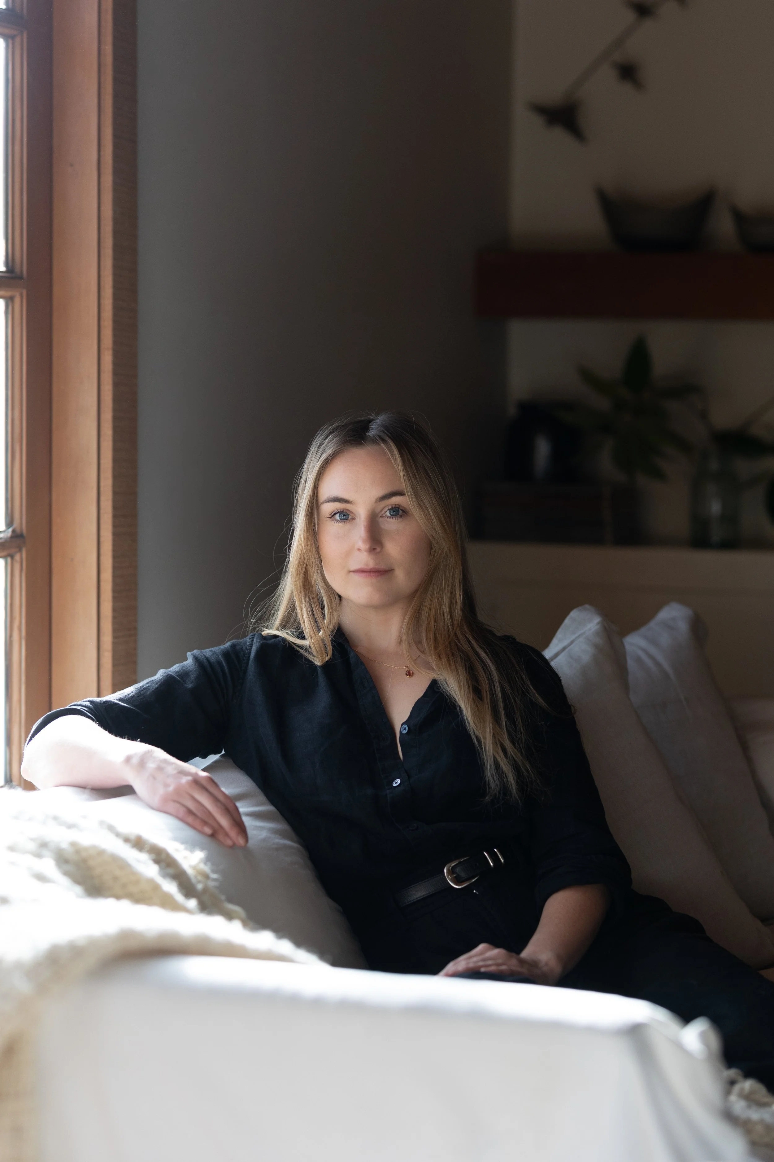 A woman with long blonde hair, wearing a black shirt, sitting on a beige sofa near a window, looking at the camera in a living room with dark walls and decorative items on shelves.