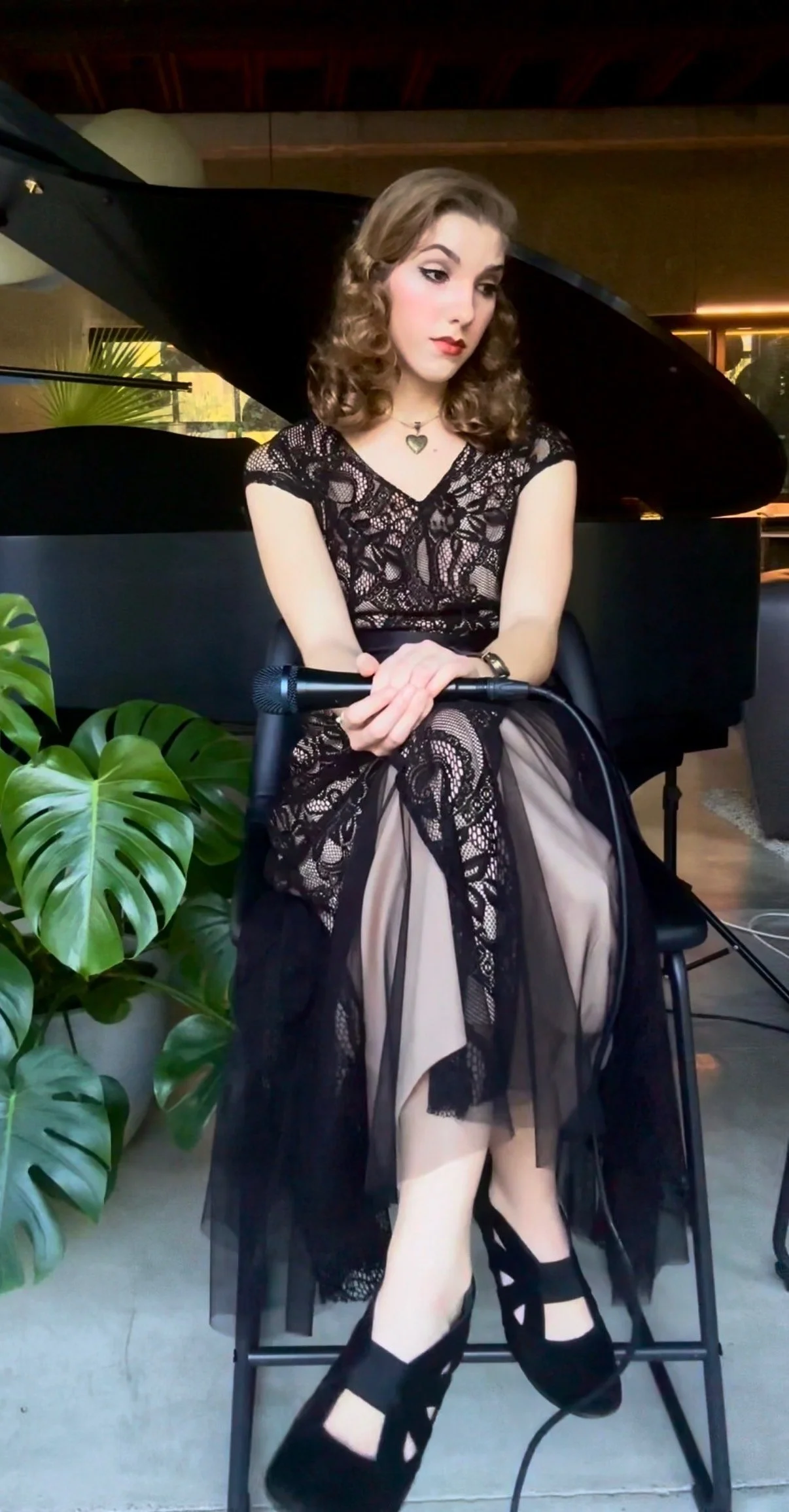A woman sitting in front of a black grand piano, holding a microphone, with a large green plant beside her. She wears a black lace dress and black high-heeled shoes.