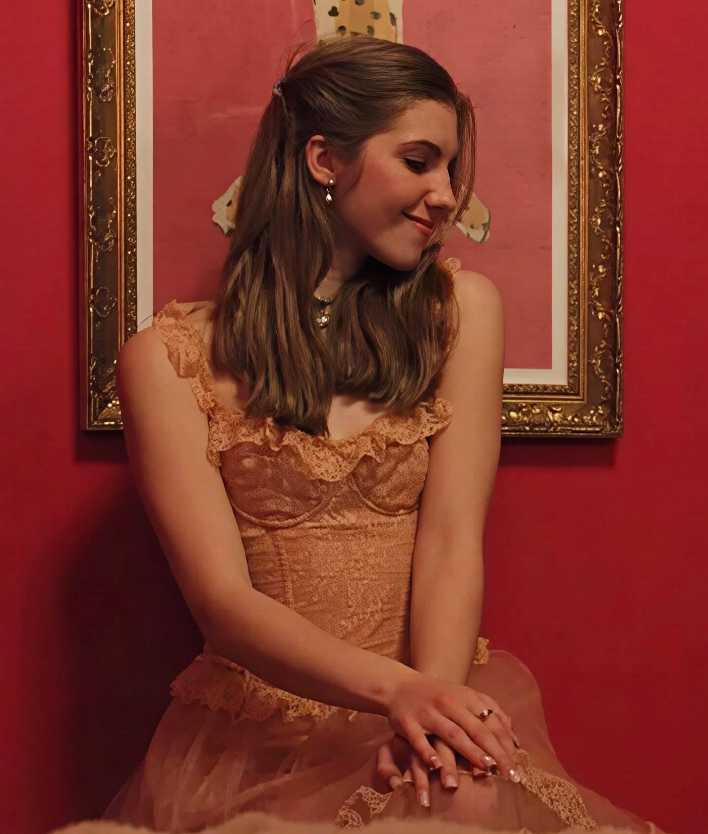 A young woman with long brown hair is sitting against a red wall with a gold-framed painting behind her. She is wearing a peach-colored lace dress and has her hands resting on her lap, with her head slightly tilted and eyes closed, smiling gently.