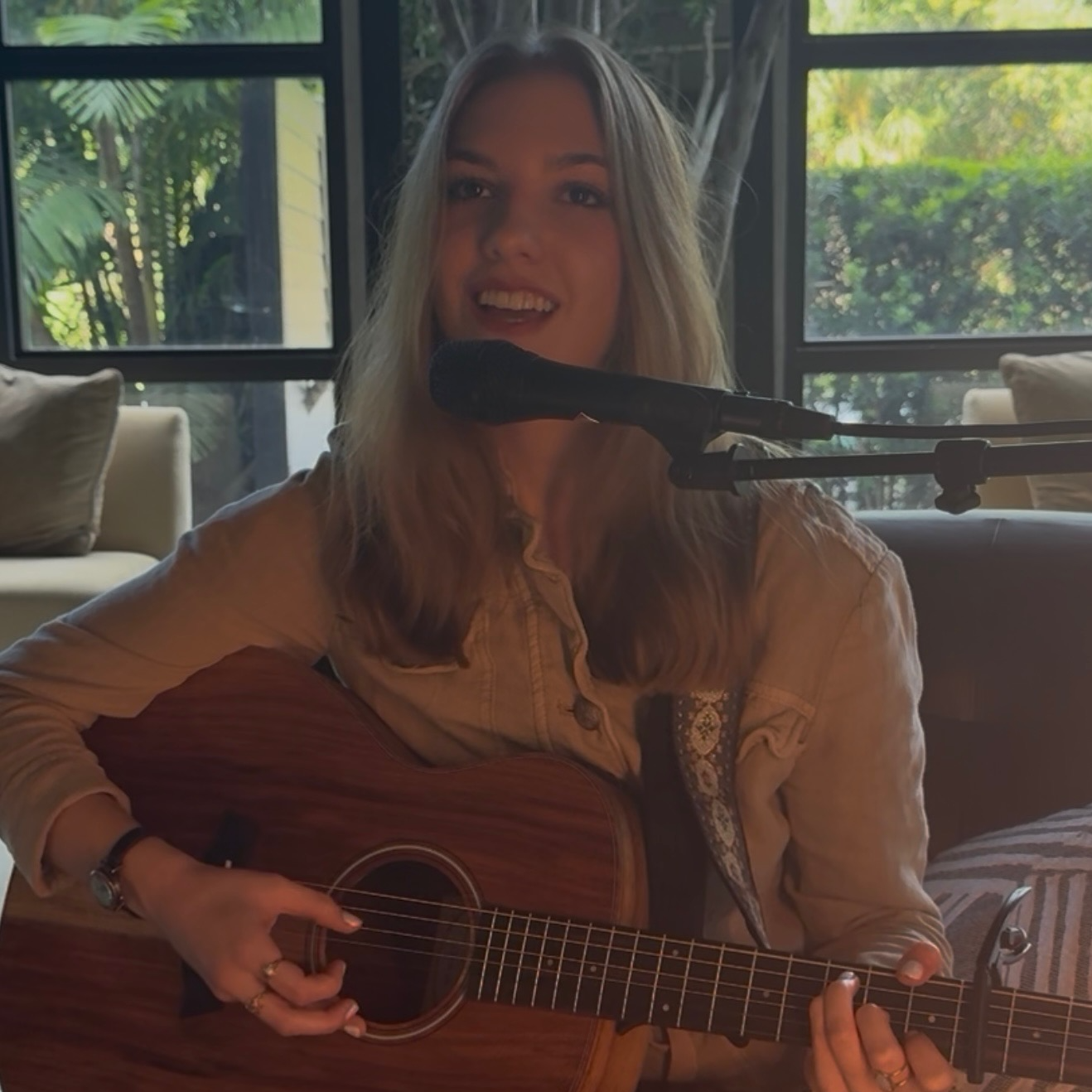 Woman singing into a microphone while playing an acoustic guitar in a room with large windows and green outdoor scenery.