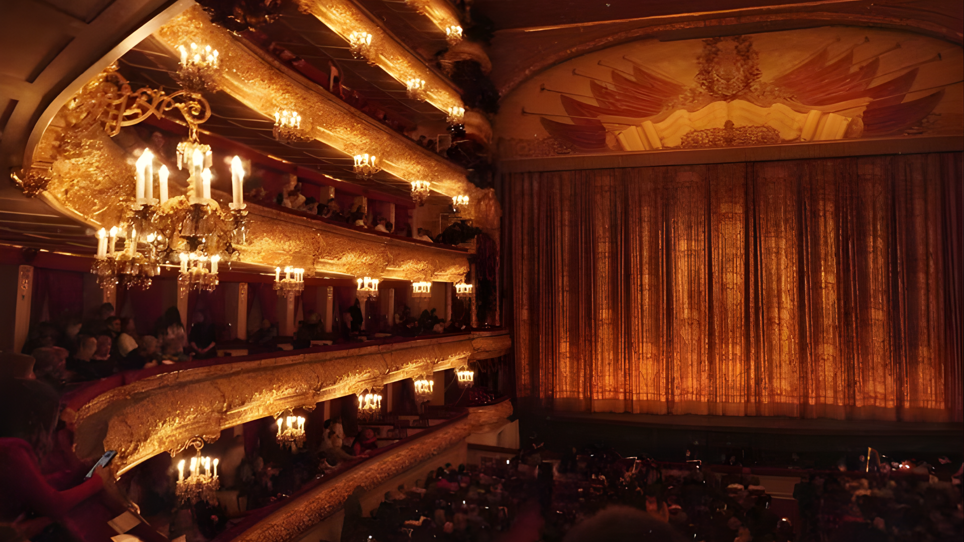 Interior view of a grand theater or opera house with ornate gold balconies, chandeliers, and a large closed curtain on the stage.