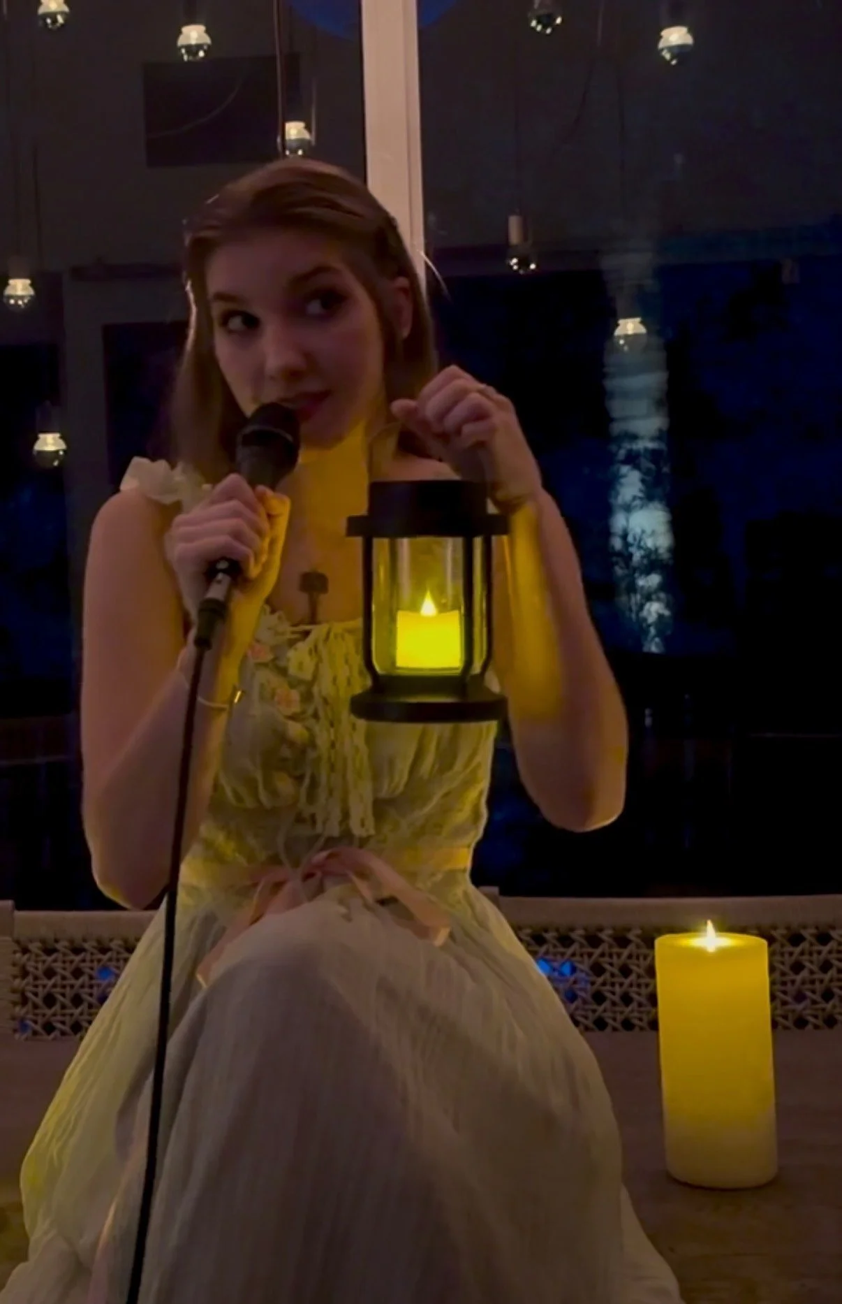 A young woman in a yellow dress sitting near lit candles, holding a microphone, with dark background and string lights overhead.