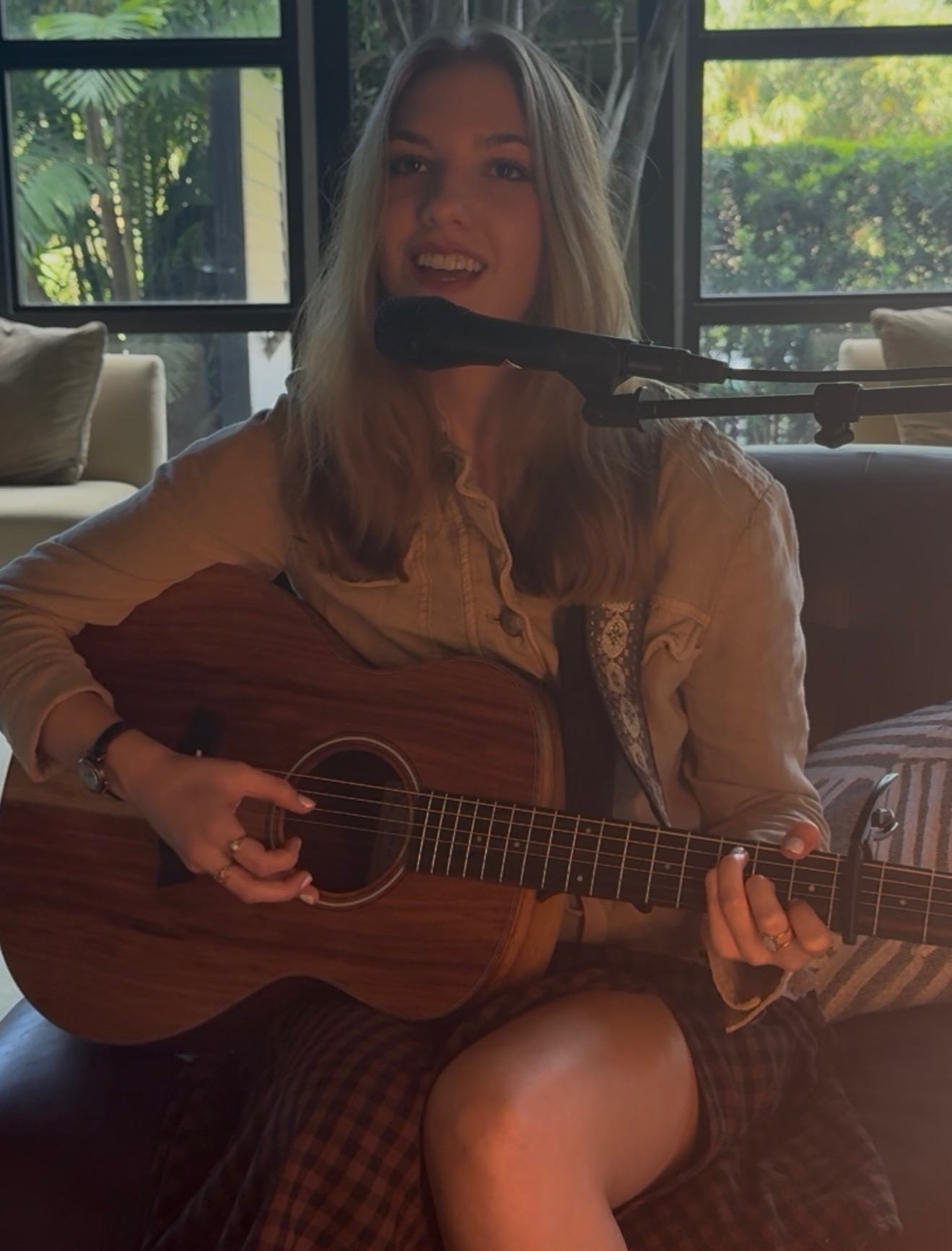 Young woman playing an acoustic guitar and singing into a microphone indoors, with windows and greenery visible in the background.