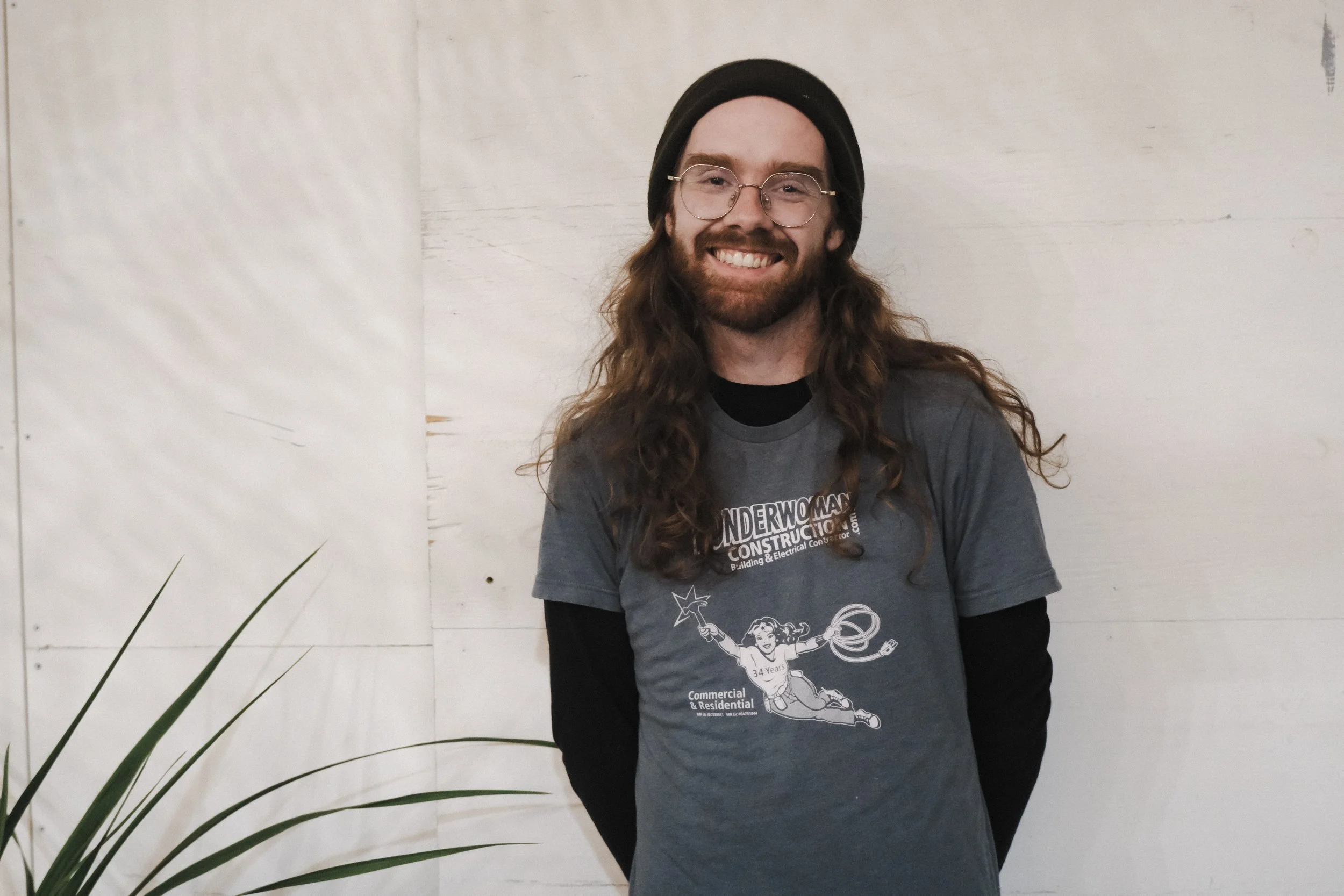 A man with long, wavy hair, a beard, and glasses, smiling in front of a white wall, wearing a black beanie and a gray t-shirt with a graphic of a girl jumping with a wand and the text 'Underground Construction'.