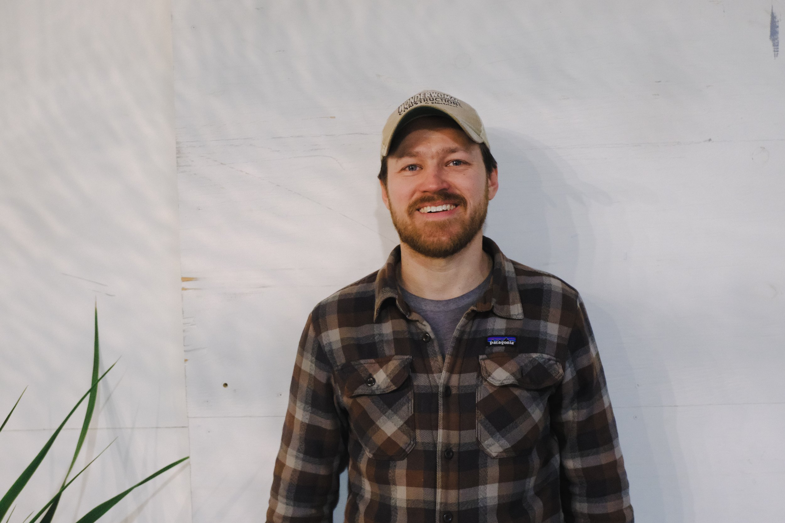 A man with a beard and mustache wearing a beige cap and a brown plaid shirt stands in front of a white wall, smiling at the camera.