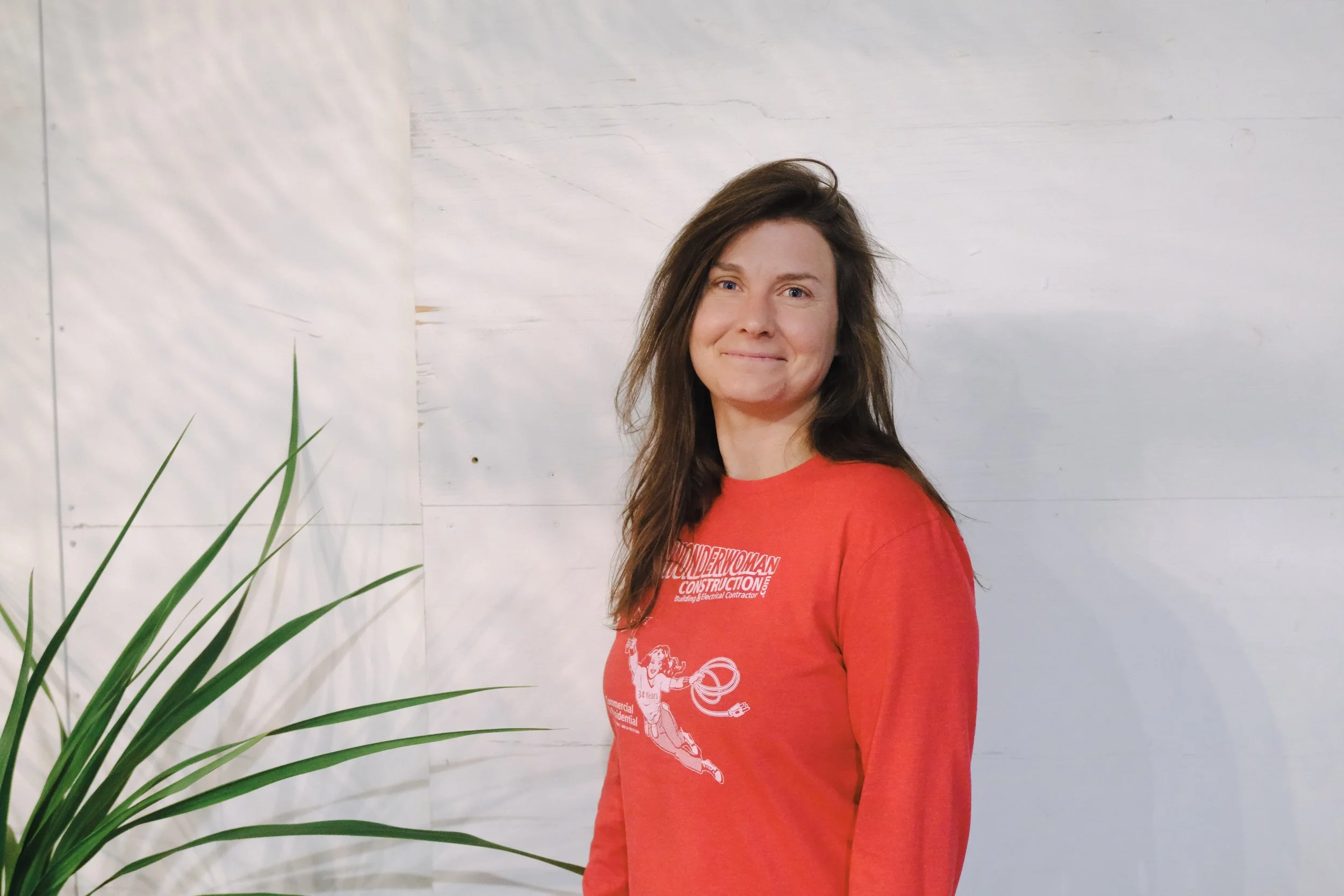Woman with long brown hair wearing a red sweatshirt with text and a graphic, standing outdoors near a white wooden wall and a green plant.