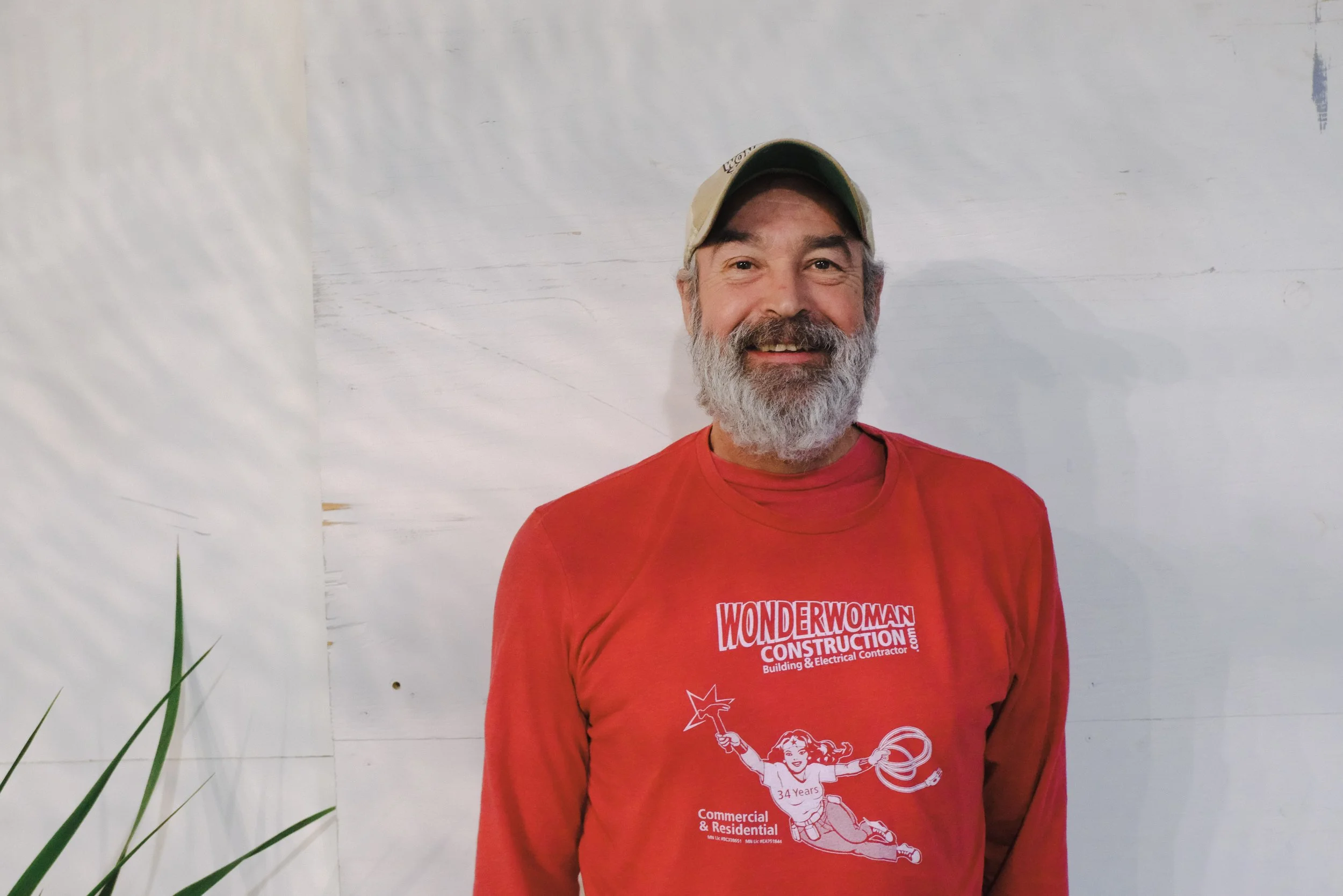 A smiling man with a beard wearing a beige cap and a red shirt with a Wonder Woman graphic and text, standing in front of a light-colored wall.