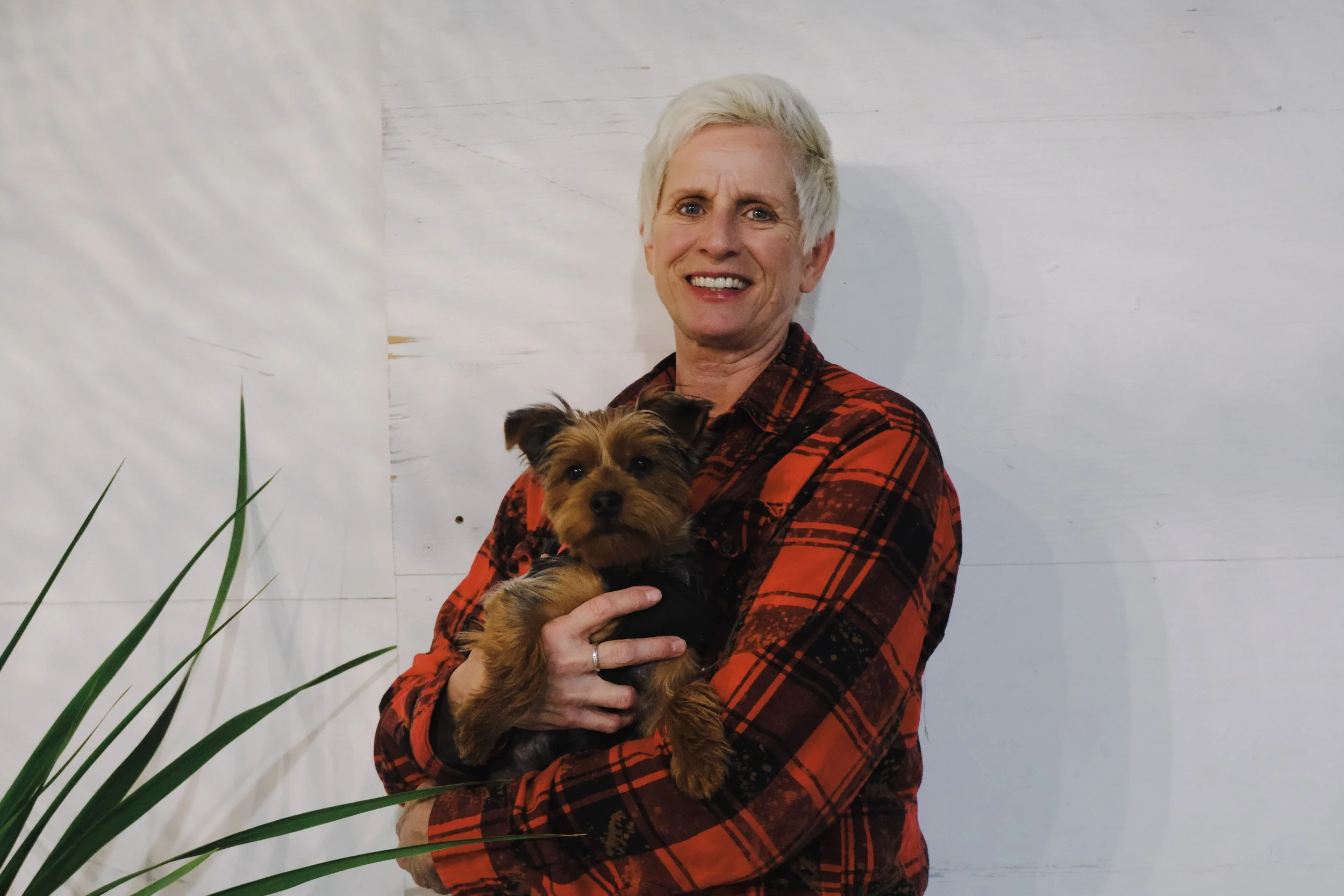A woman with short gray hair wearing a red and black plaid shirt holding a small brown dog with black fur on its back.