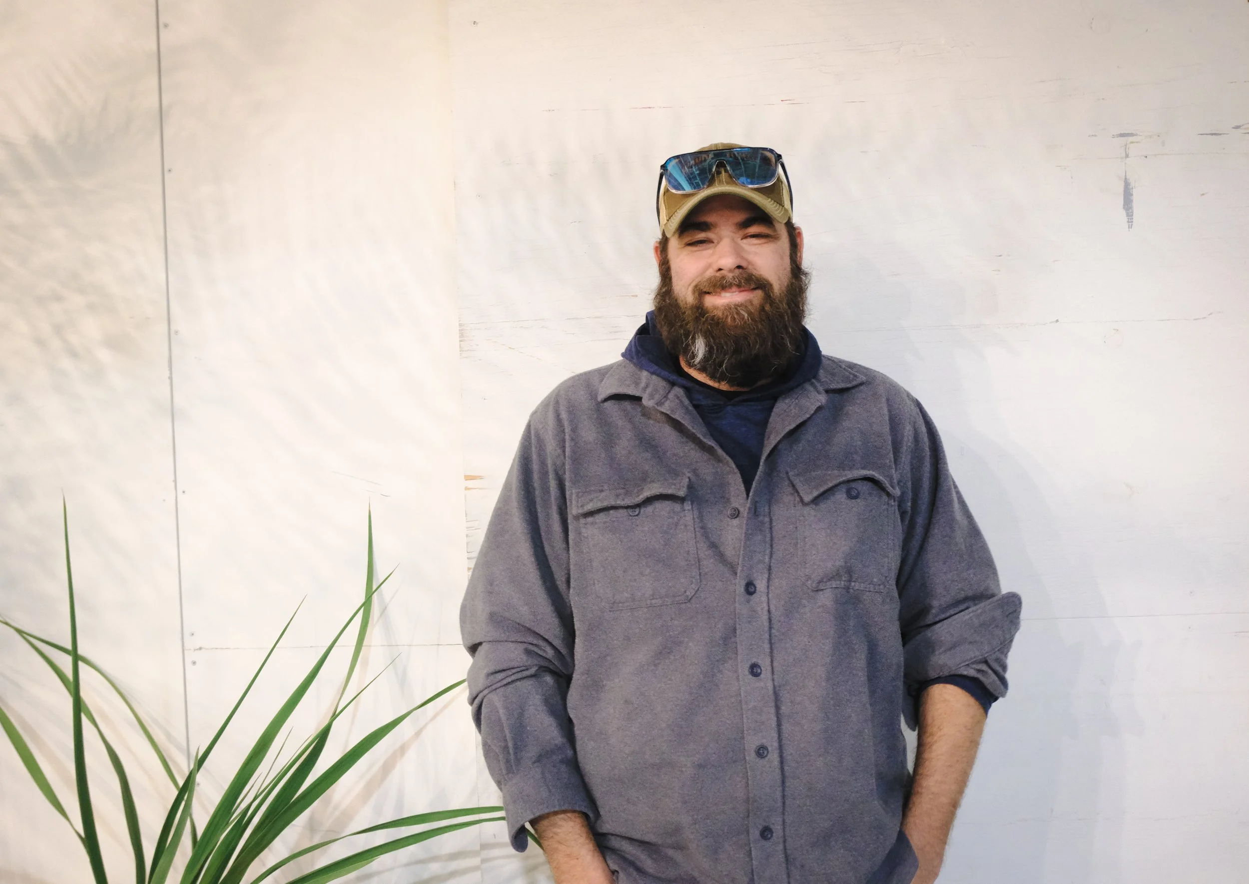 A man with a beard, wearing a baseball cap and sunglasses on his head, standing in front of a white wall with a plant to the left.