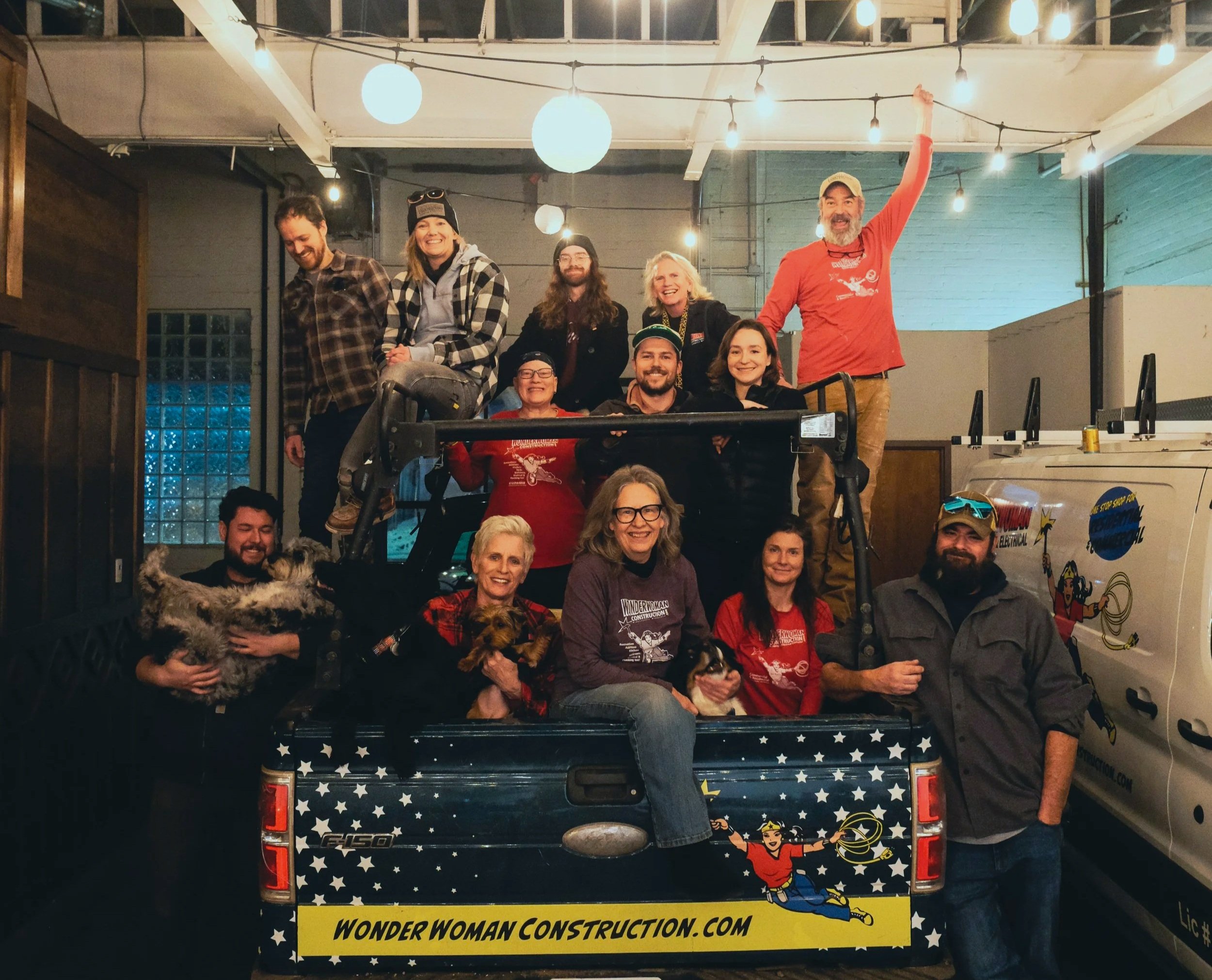 Group of people in a decorated pickup truck with a Wonder Woman theme, inside a room with hanging lights. Some individuals are holding dogs, and the truck has a banner with a Wonder Woman illustration and the website wonderwomanconstruction.com.
