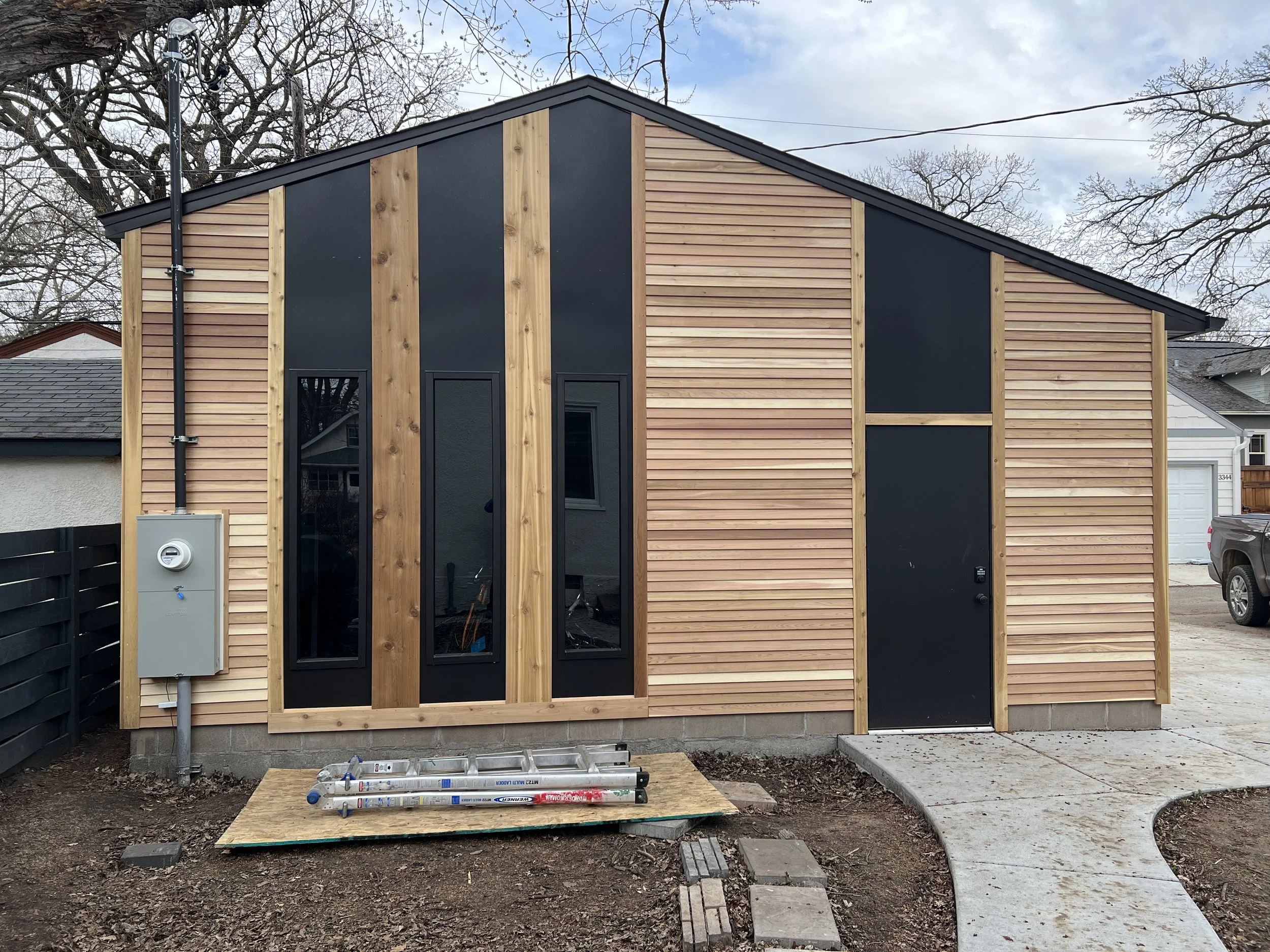 Newly constructed small building with vertical wood siding and black accents, with a curved concrete walkway leading to a black door, bare trees in the background, and construction tools on the ground in front.
