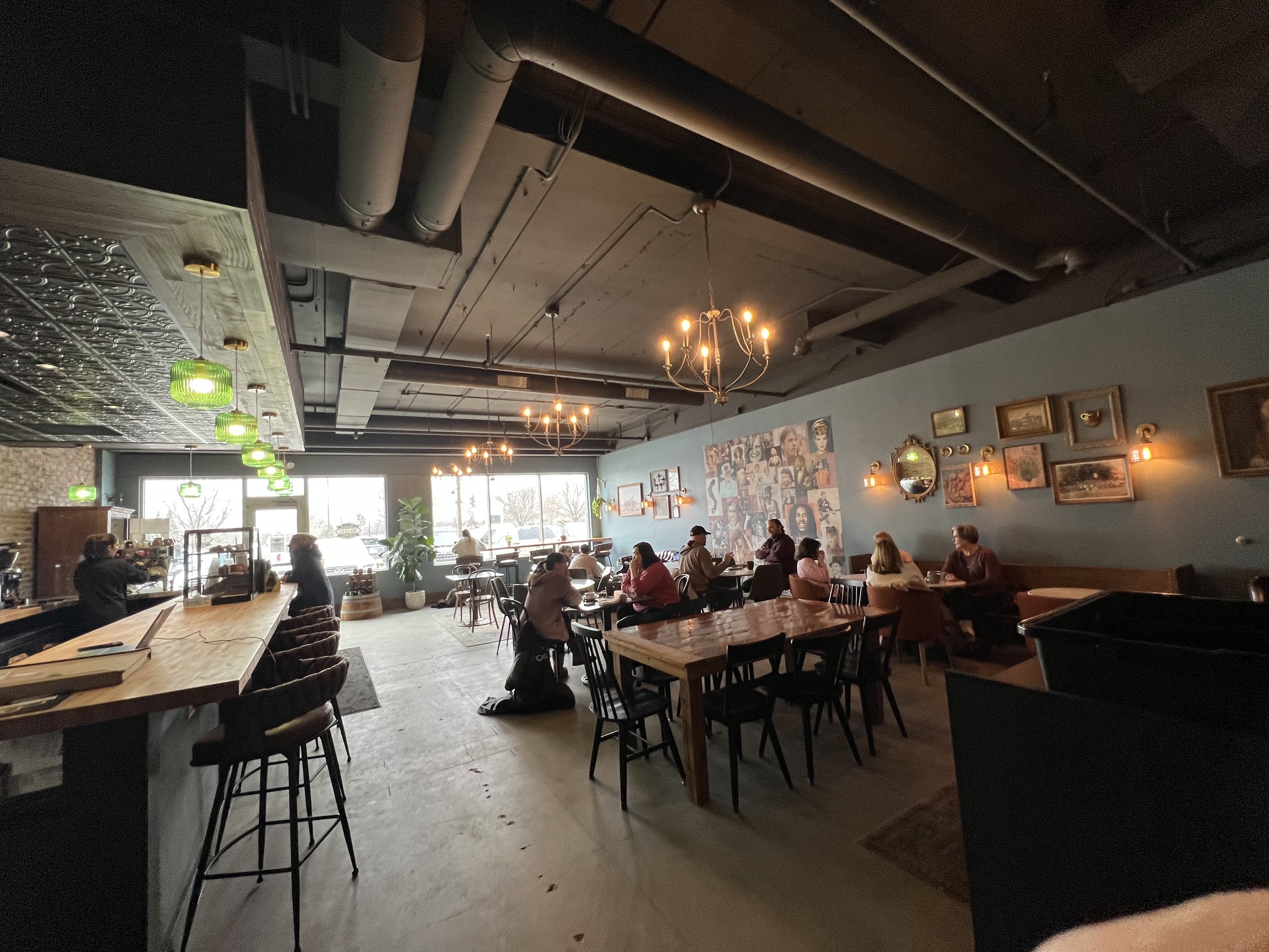Interior of a cozy coffee shop with customers sitting at tables, a barista working at the counter, vintage artwork and mirrors on the walls, chandeliers and green pendant lights hanging from a dark ceiling, and large windows letting in natural light.