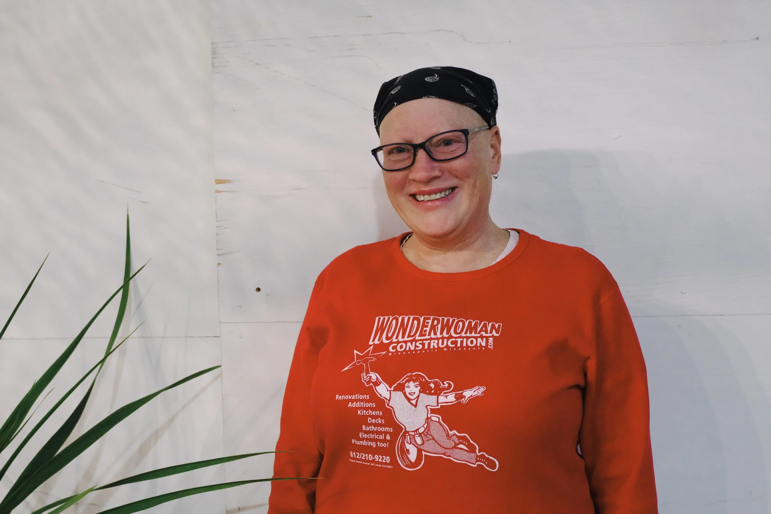 A woman with glasses and a bandana, smiling, wearing an orange Wonder Woman construction company sweatshirt, standing in front of a white textured wall with a plant in the foreground.