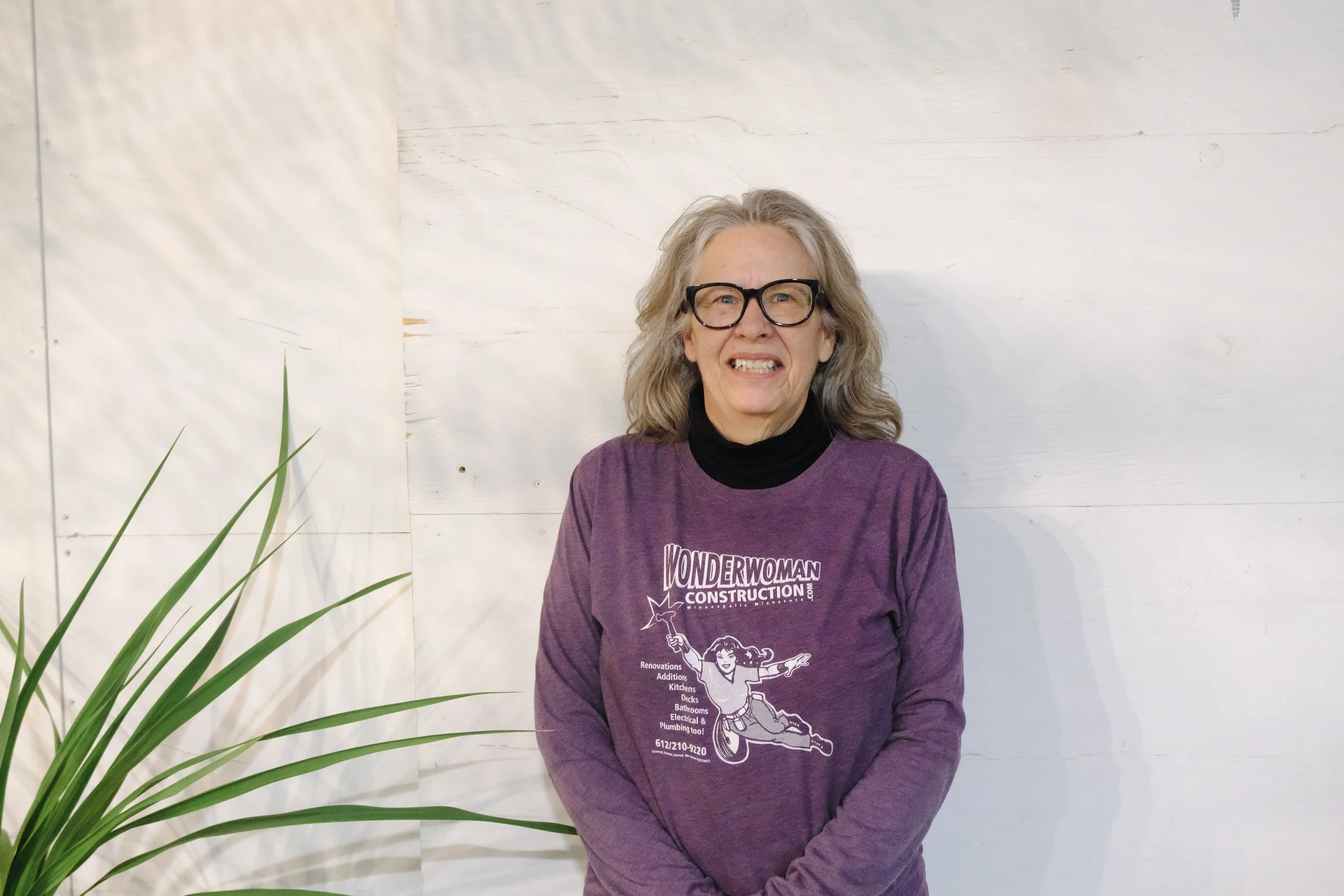A woman with gray hair and glasses wearing a purple Wonder Woman Construction t-shirt, standing in front of a white wooden wall.