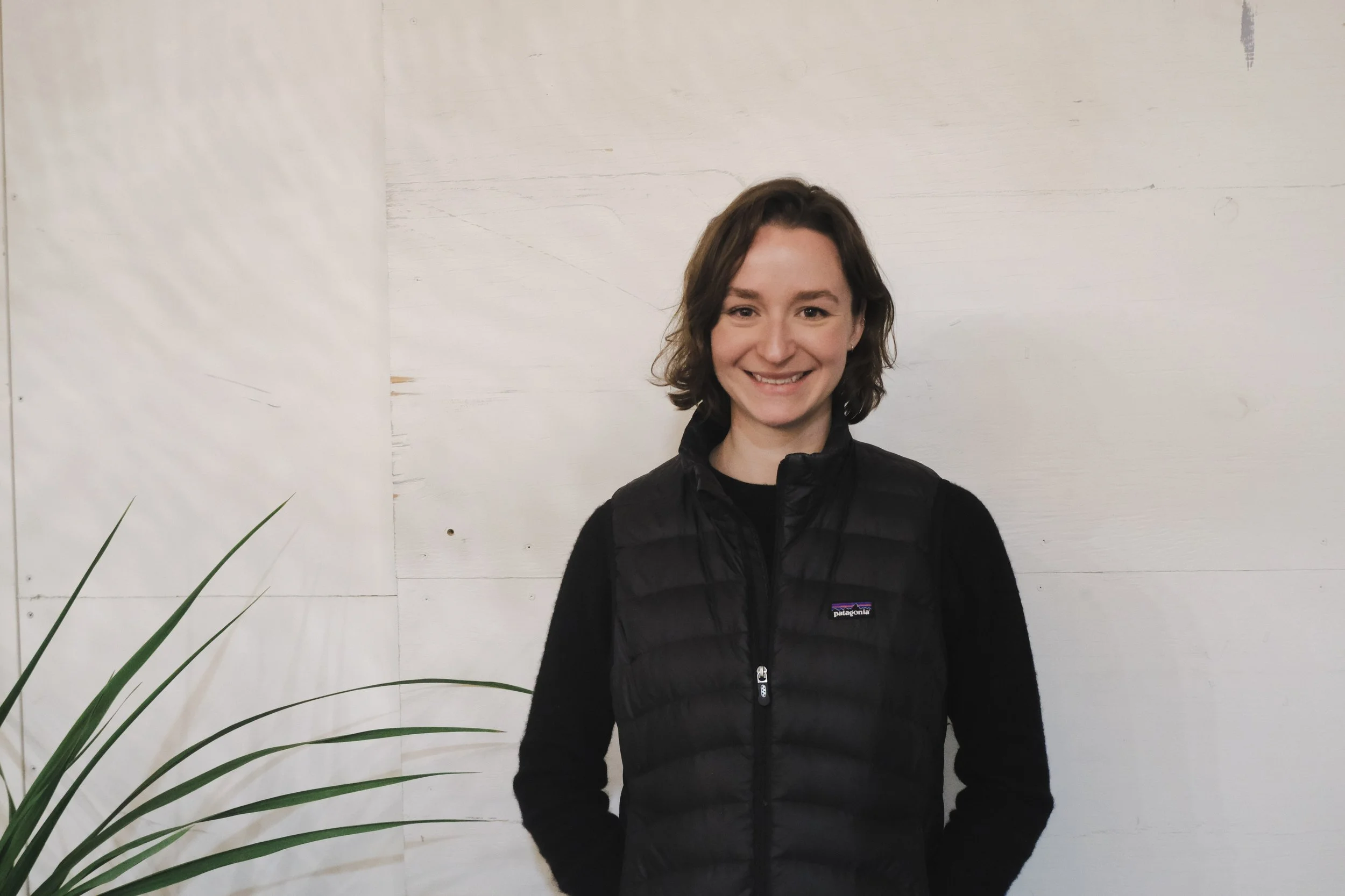A woman with shoulder-length brown hair smiling, wearing a black Patagonia puffer vest over a black long sleeve shirt, standing in front of a white wooden wall with some green plant leaves visible in the lower left corner.