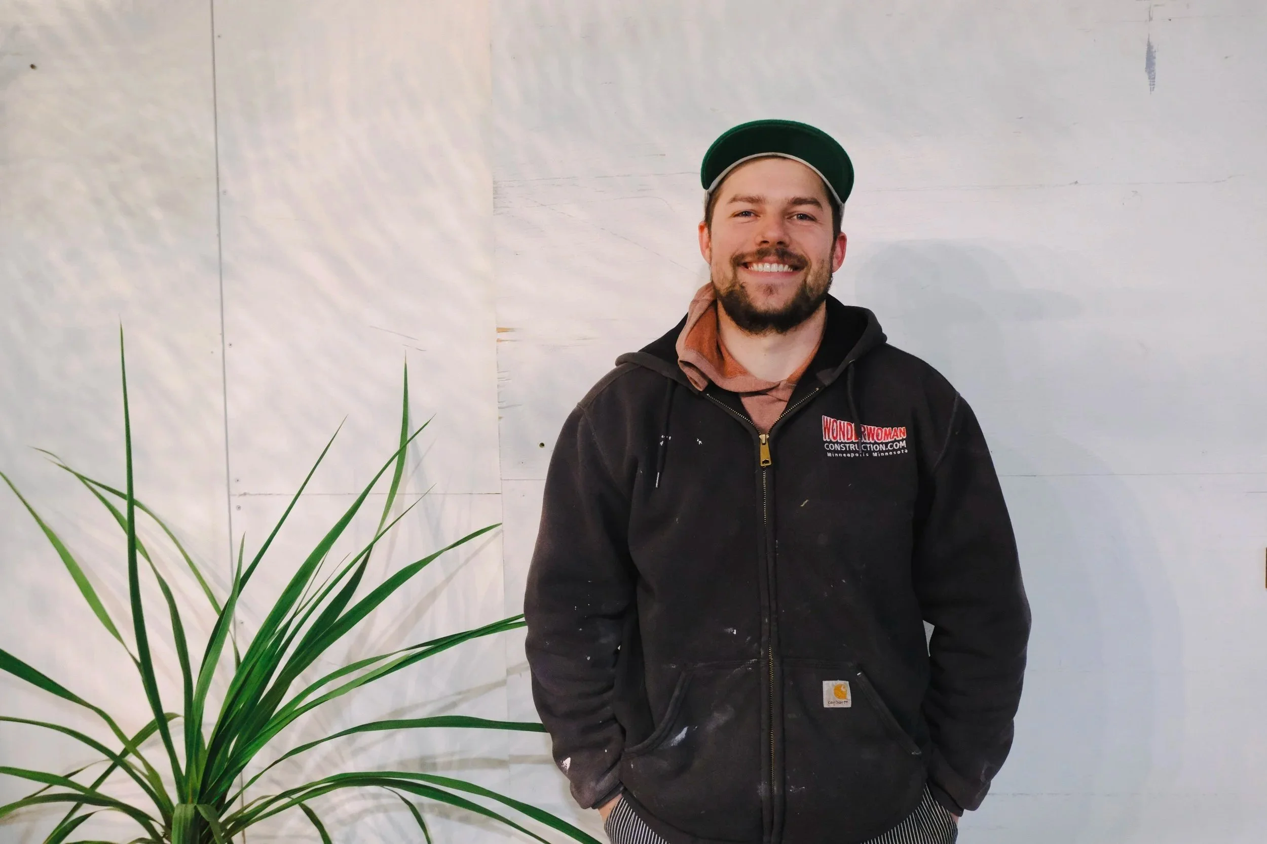 A smiling man with a beard, wearing a green cap, a black Carhartt jacket with paint stains, standing indoors next to a large green plant. The background is a plain white wall.