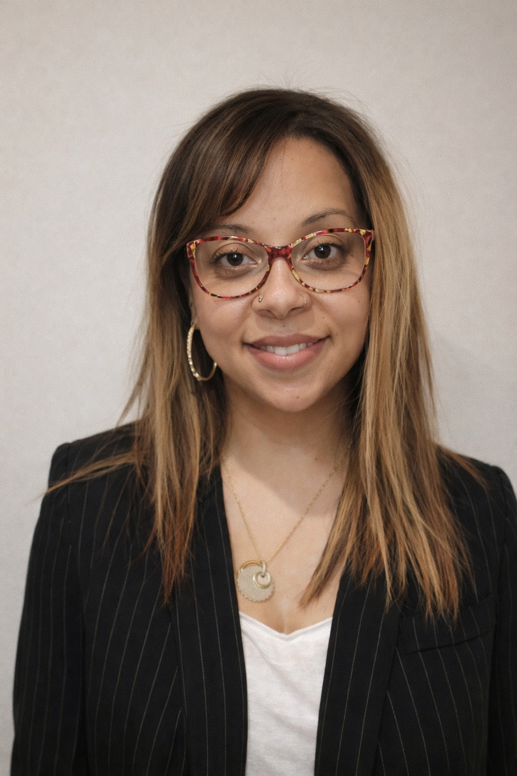 A young woman with light brown hair, wearing red and gold tortoiseshell glasses, hoop earrings, a gold necklace, and a black pinstripe blazer, smiling in front of a plain white background.