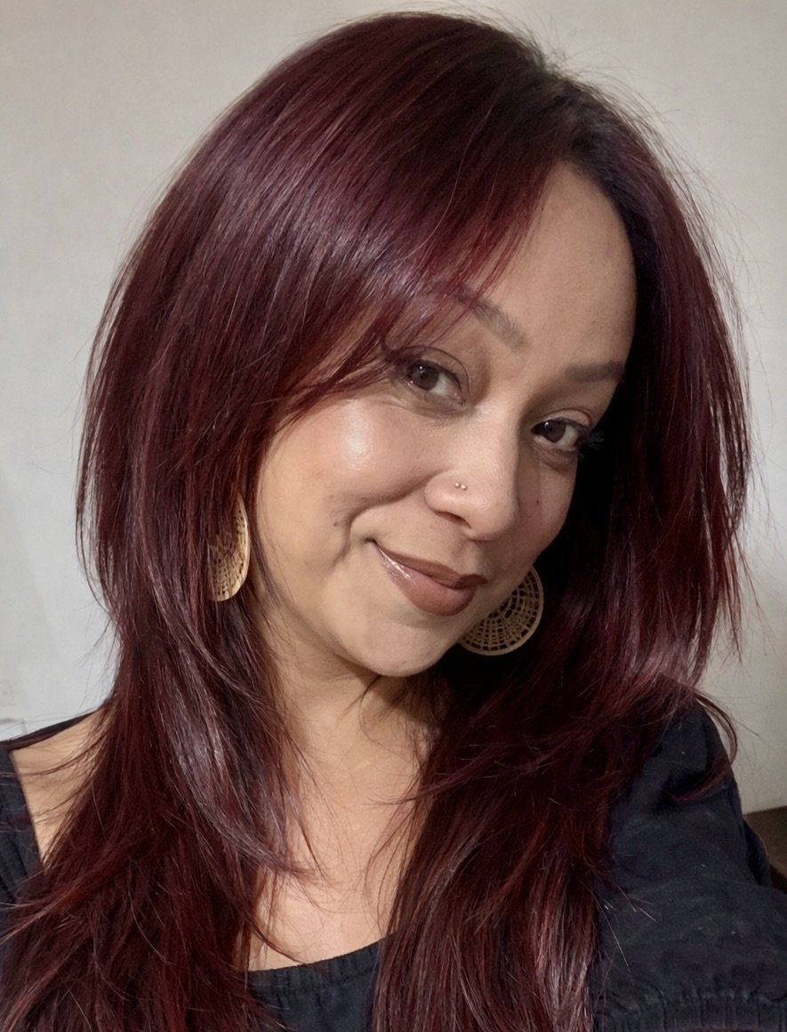 A woman with shoulder-length, reddish-brown hair, wearing large round earrings and a black top, smiling softly at the camera.
