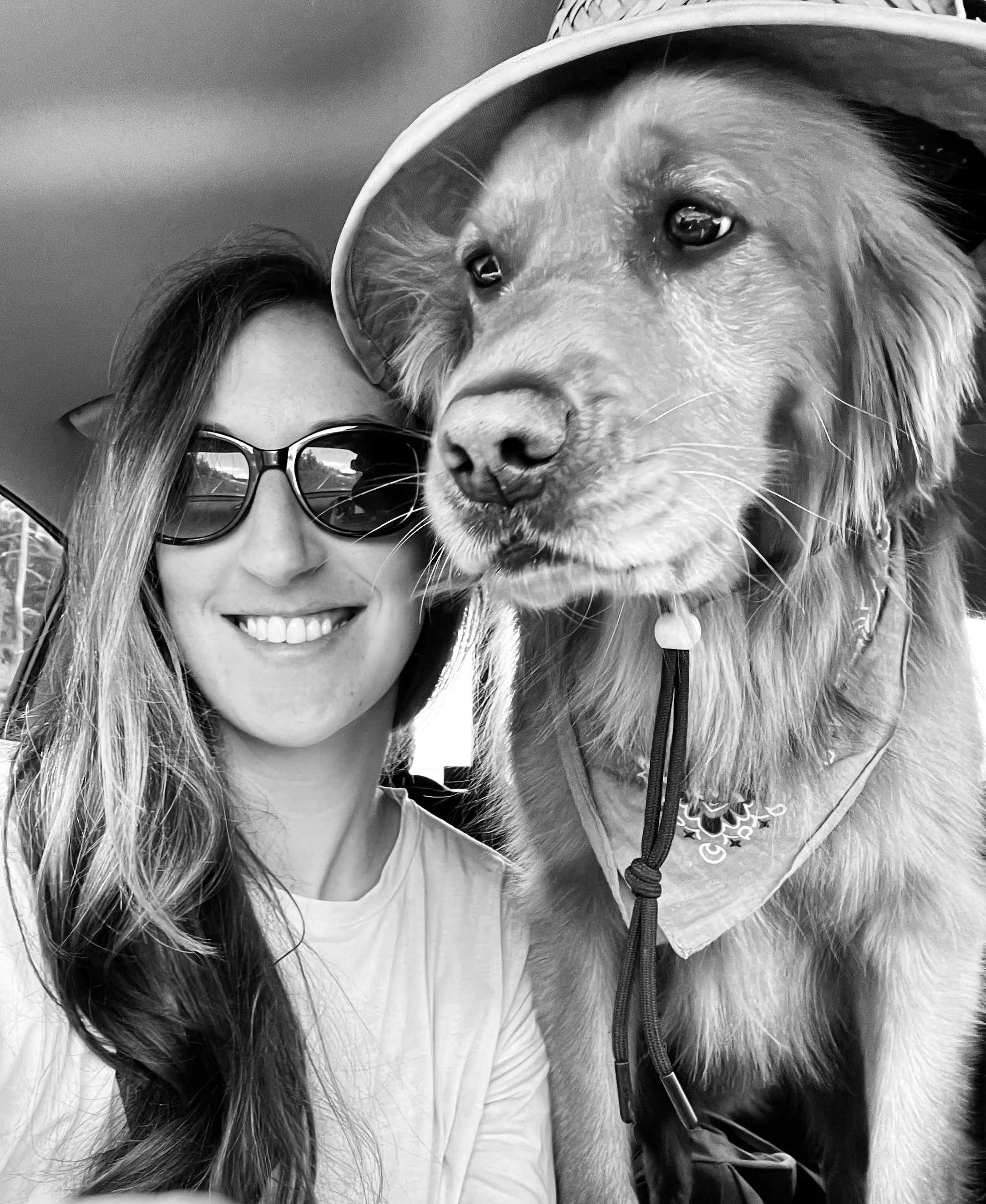 A woman with long hair and sunglasses smiling next to a large Golden Retriever wearing a hat and bandana inside a vehicle.