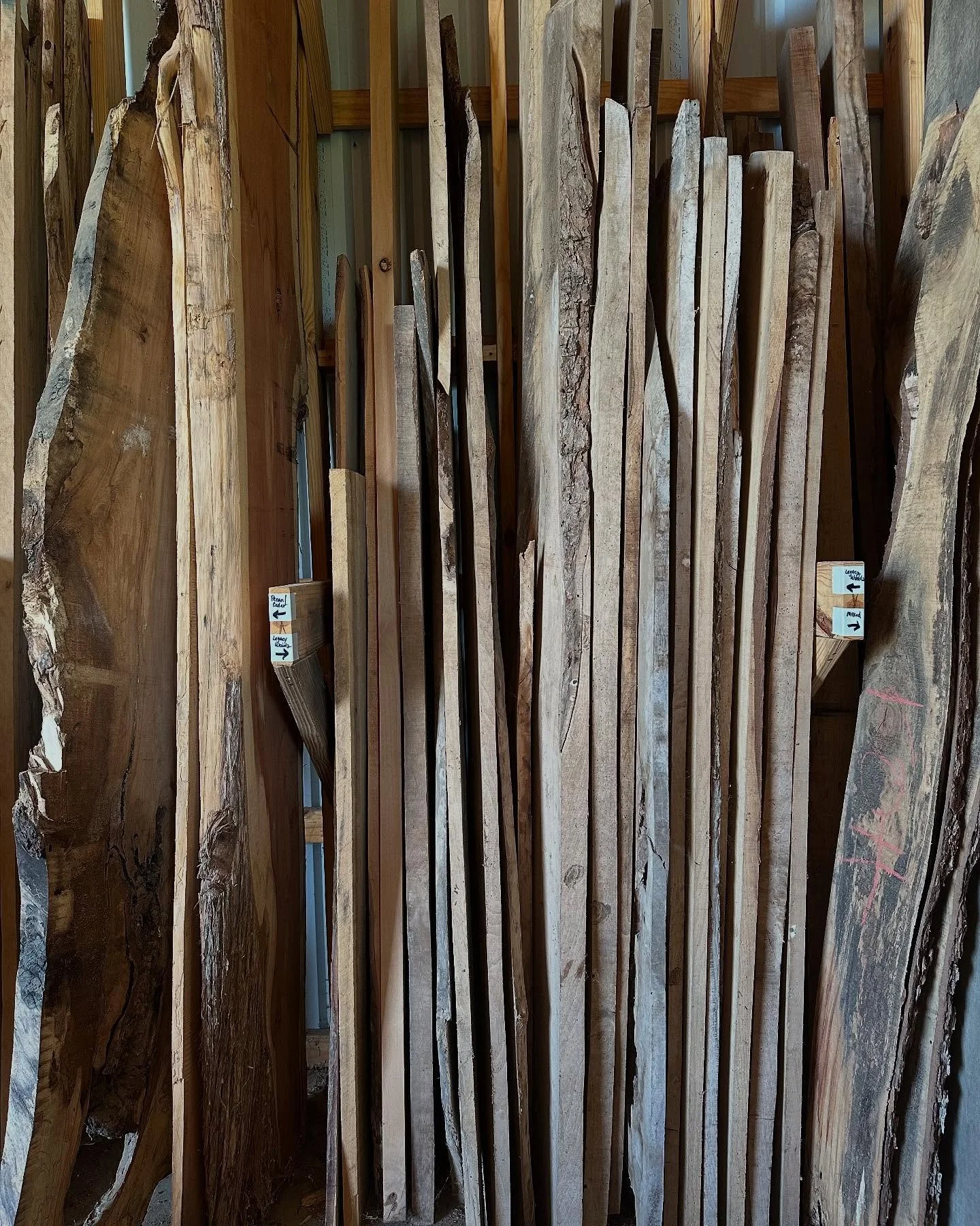 Various lengths and types of natural wood planks and slabs stored vertically in a woodworking shop, with small tags attached to some pieces.