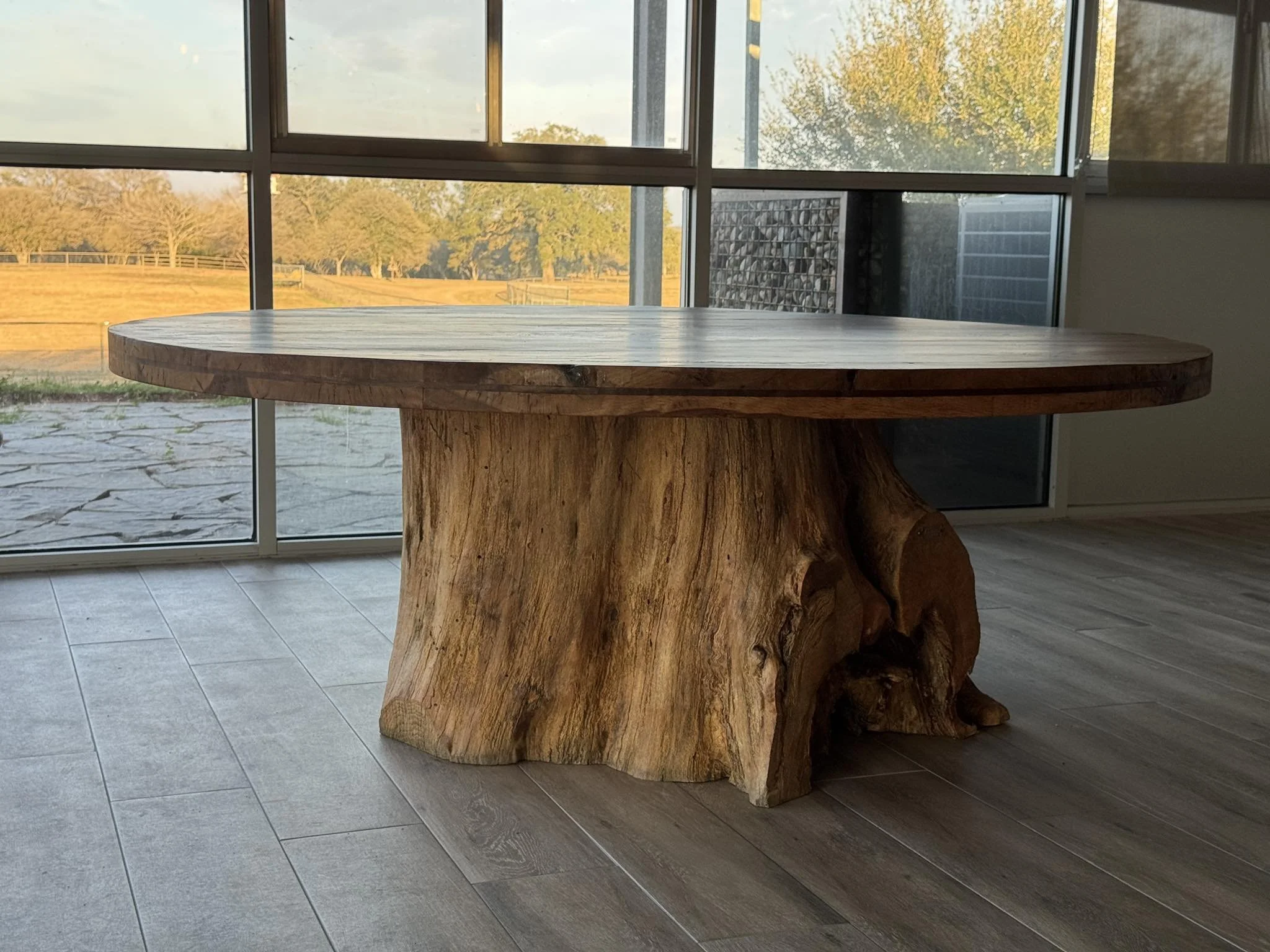 A round wooden table with a natural, rustic finish, standing on a large, thick tree stump base inside a room with large windows showing an outdoor landscape with trees and a field at sunset.