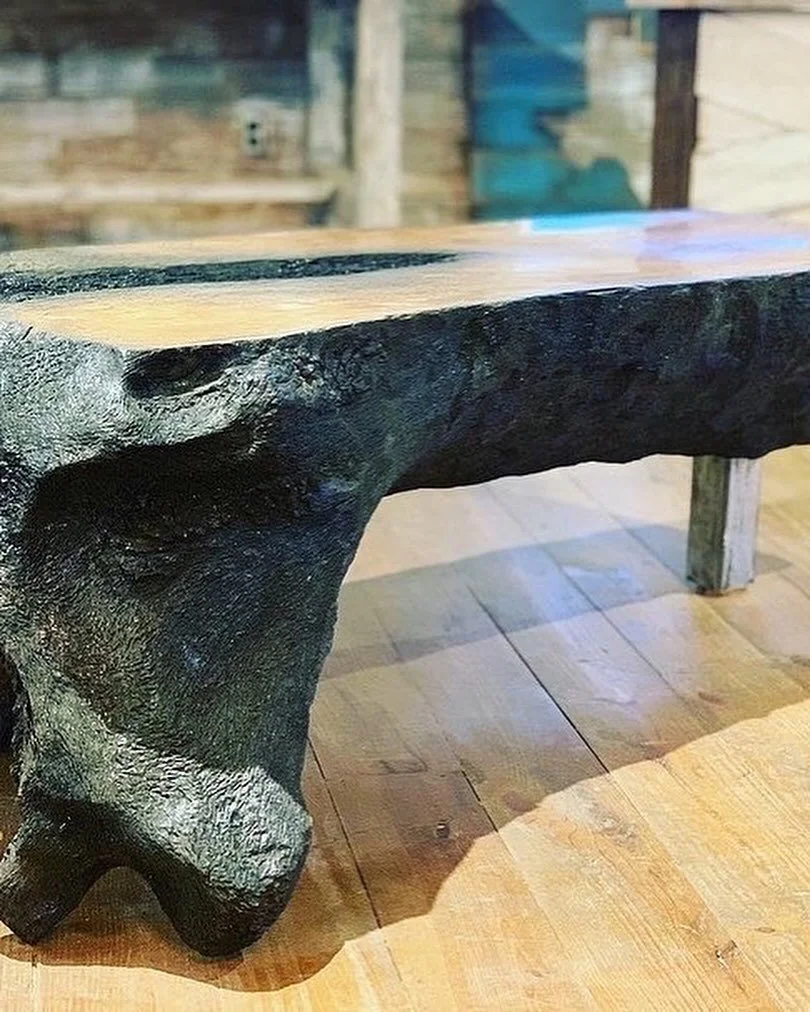 Close-up of a wooden table with a black, textured, tree trunk-shaped bench in an indoor setting with wooden flooring and a blurred background.