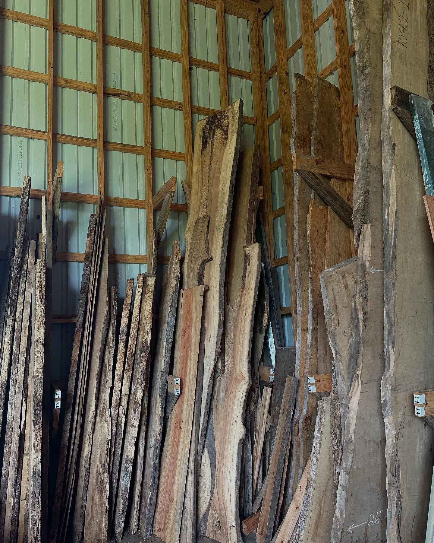 Stack of raw, natural wood planks and slabs leaning against a metal wall in a lumber or woodworking shop.