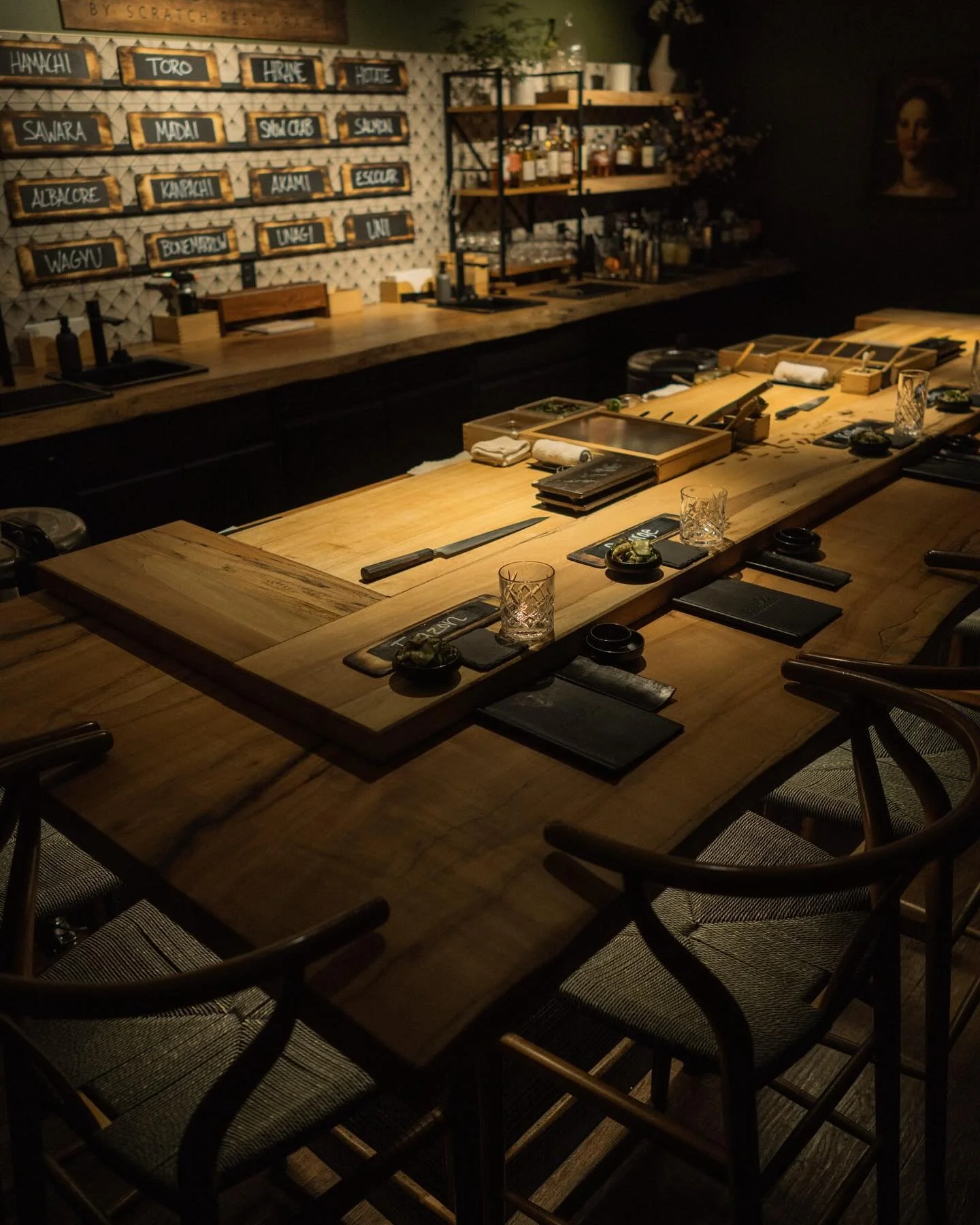 A dimly lit Japanese restaurant with a long wooden table, place settings including black plates and textured glasses, and a chalkboard wall displaying different types of fish in Japanese.