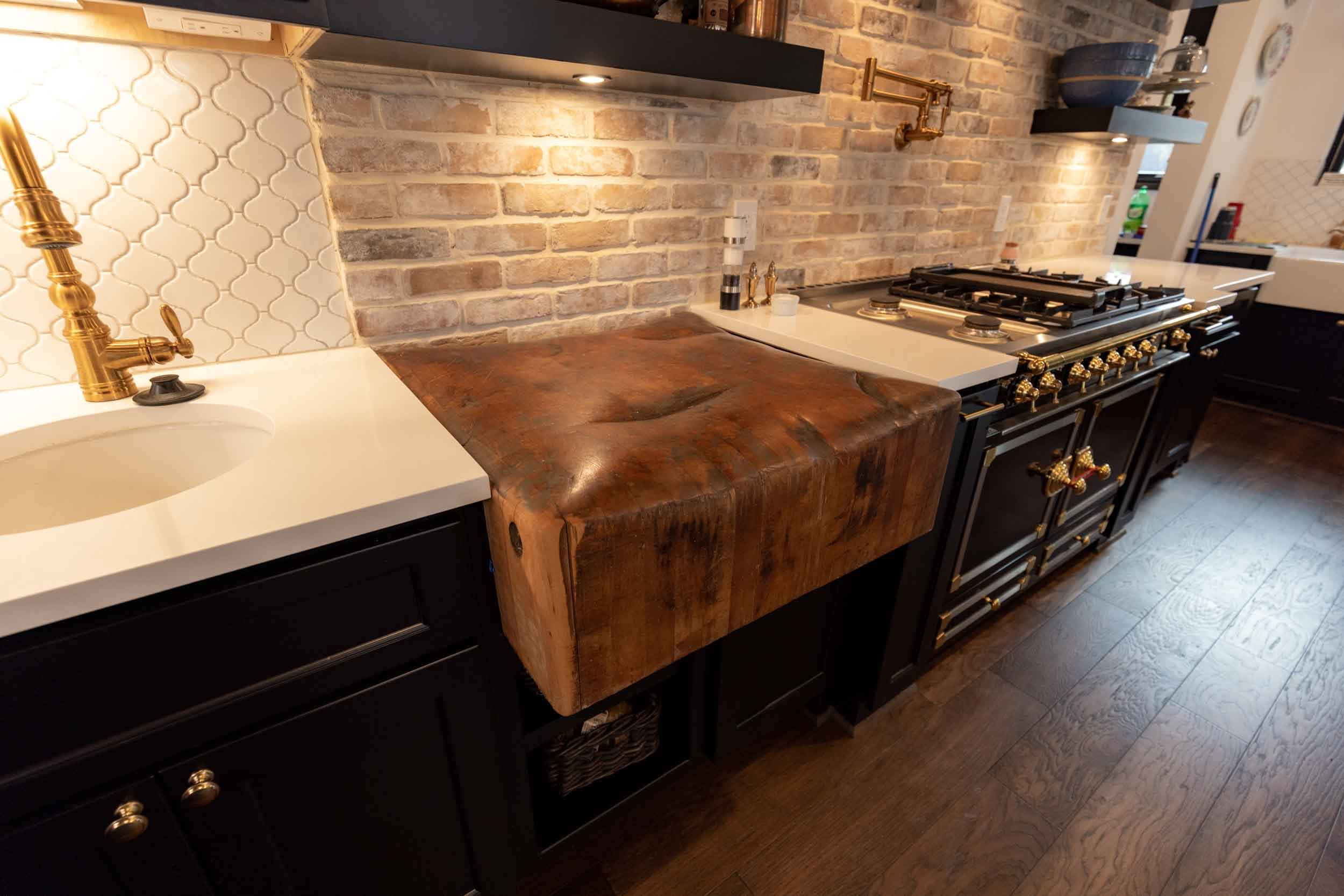 Kitchen with black cabinets, a white countertop, a brass faucet, exposed brick wall, a large rustic wooden butcher block counter, a gas stove with brass accents, and decorative shelving.