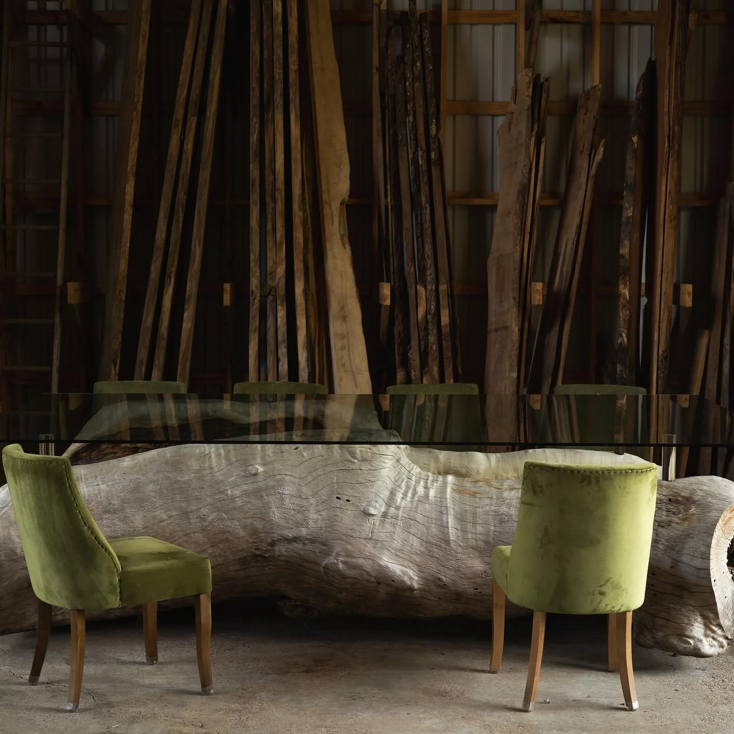 Interior space with a large rustic wooden table made from a tree trunk and two green upholstered chairs, against a background of stacked wooden planks or slats.