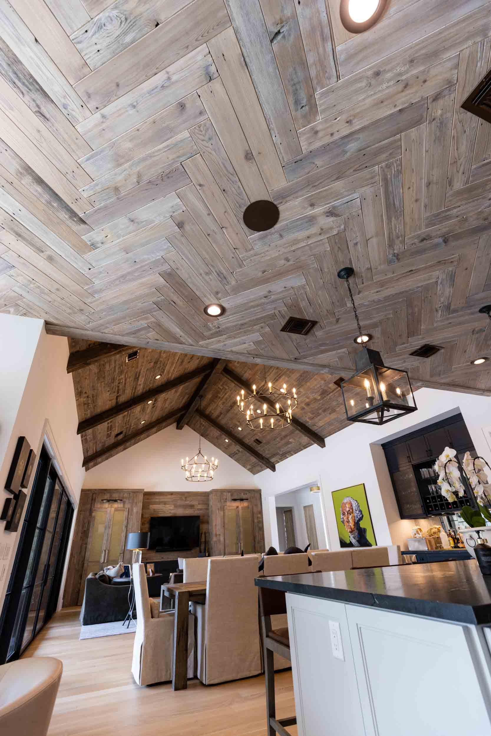Interior view of a living room with a vaulted ceiling, wooden beams, and a combination of wood and white walls. The ceiling features wood planks arranged in a herringbone pattern with recessed lighting. Two chandeliers and a pendant light hang from t