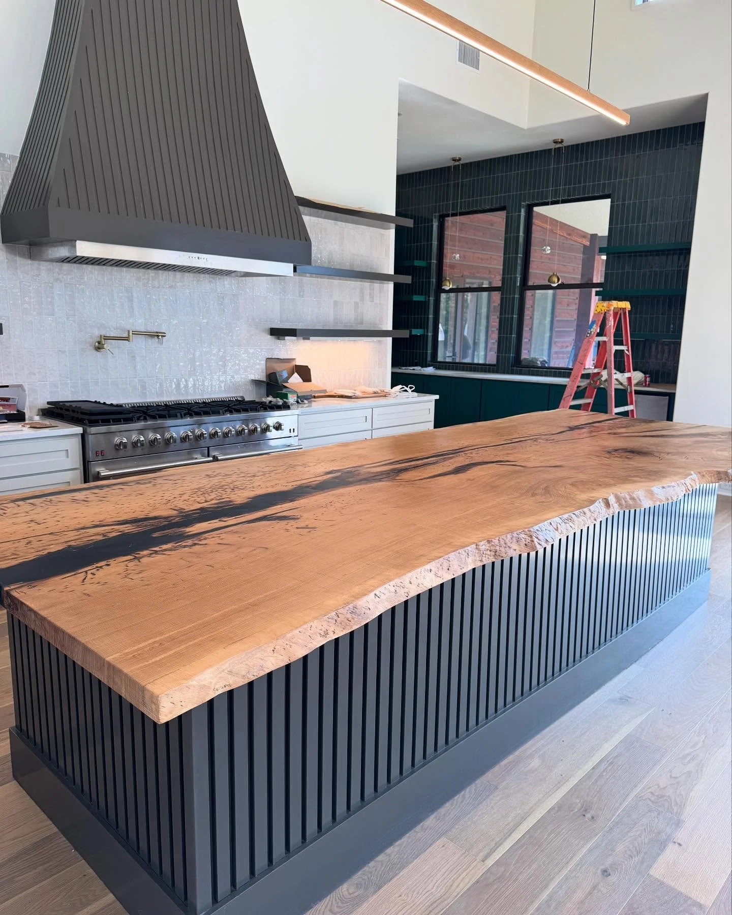 Modern kitchen with a natural wood island featuring black accents, white cabinetry, a large industrial stove, black tiled walls, and a ladder near the window.