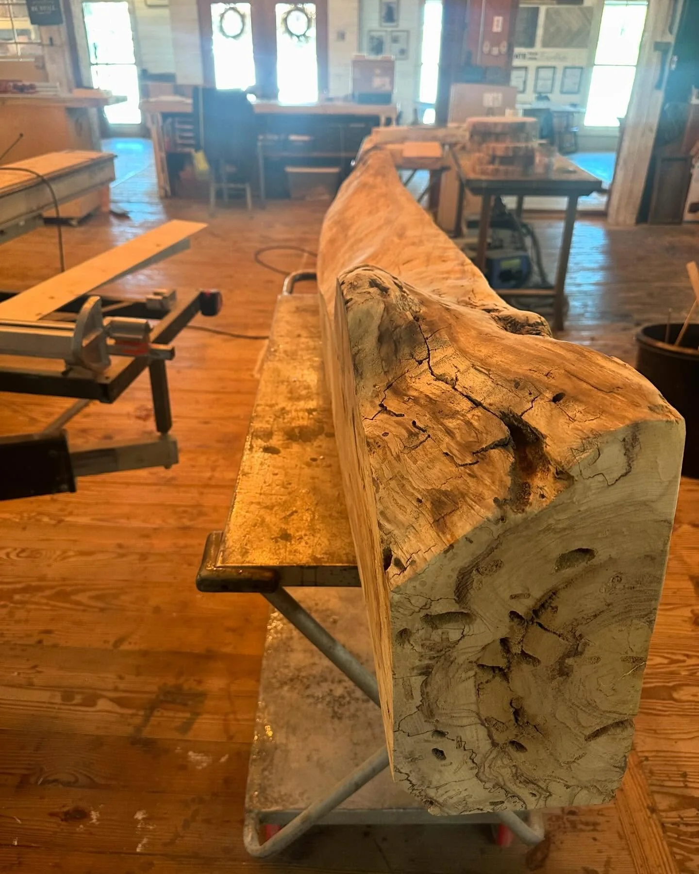 A large, rustic wooden beam with visible cracks and knots is placed on a workbench inside a woodworking shop. The shop has wooden floors, various tools, and furniture in the background.