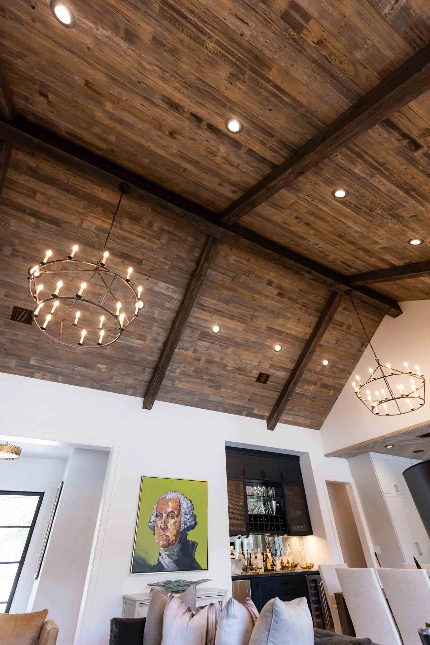 Living room with a wood-paneled vaulted ceiling, two circular chandeliers, recessed lighting, a painting of George Washington on the wall, a white cabinet, and beige chairs.