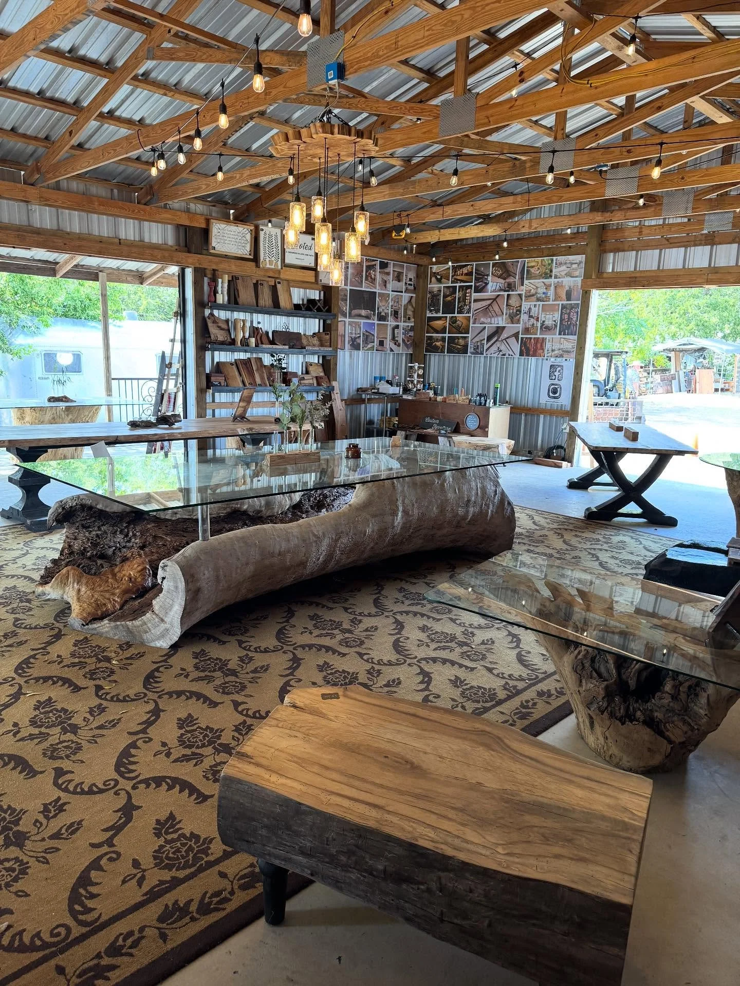 Rustic interior of a woodworking shop with wooden beams, pendant lights, and various woodworking projects and tools on display. There is a large log used as a table, a glass-topped coffee table with a wooden base, and a patterned rug on the floor.