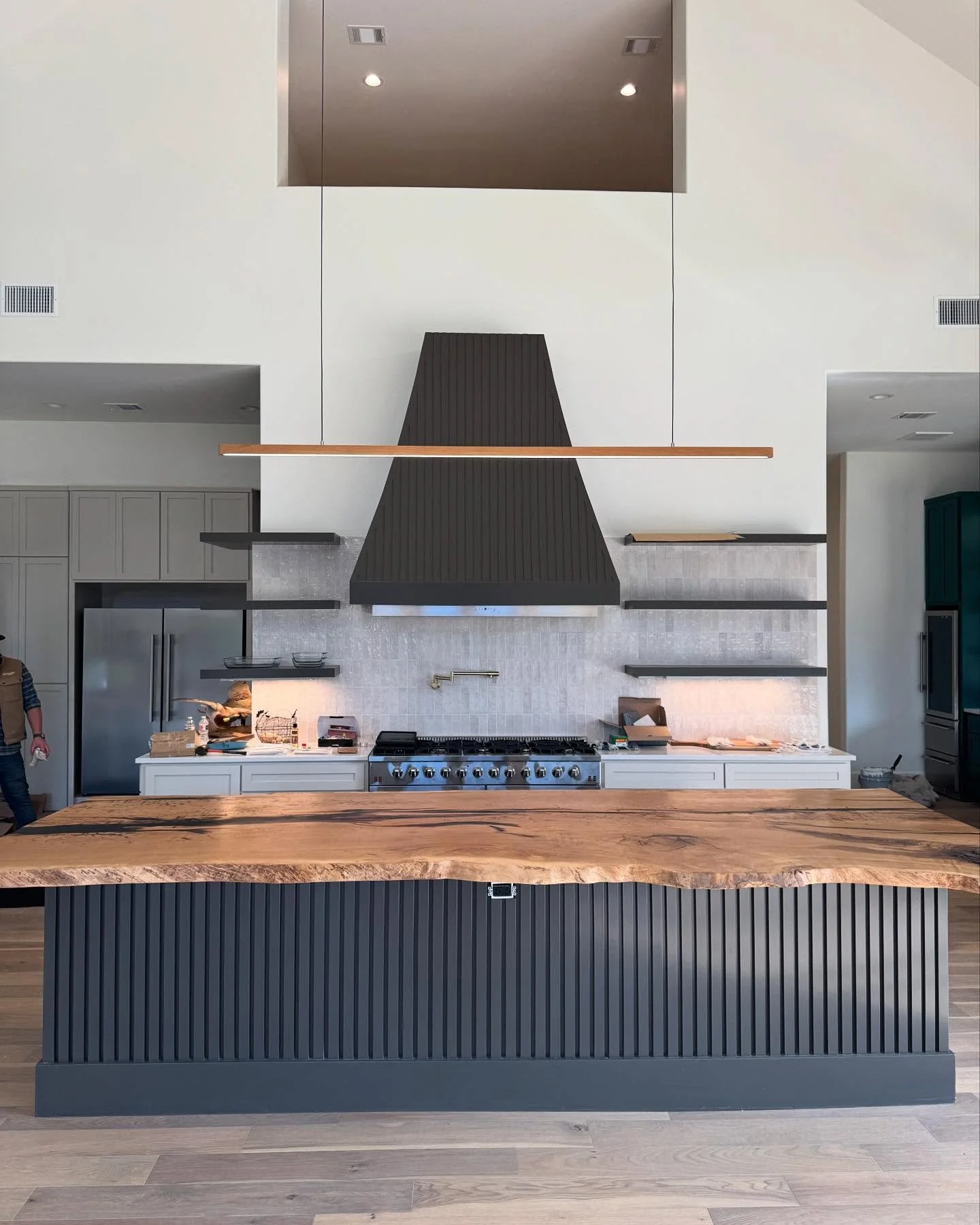 Modern kitchen with a large wooden island, black cabinetry, a black range hood, stainless steel appliances, and minimalistic open shelves.