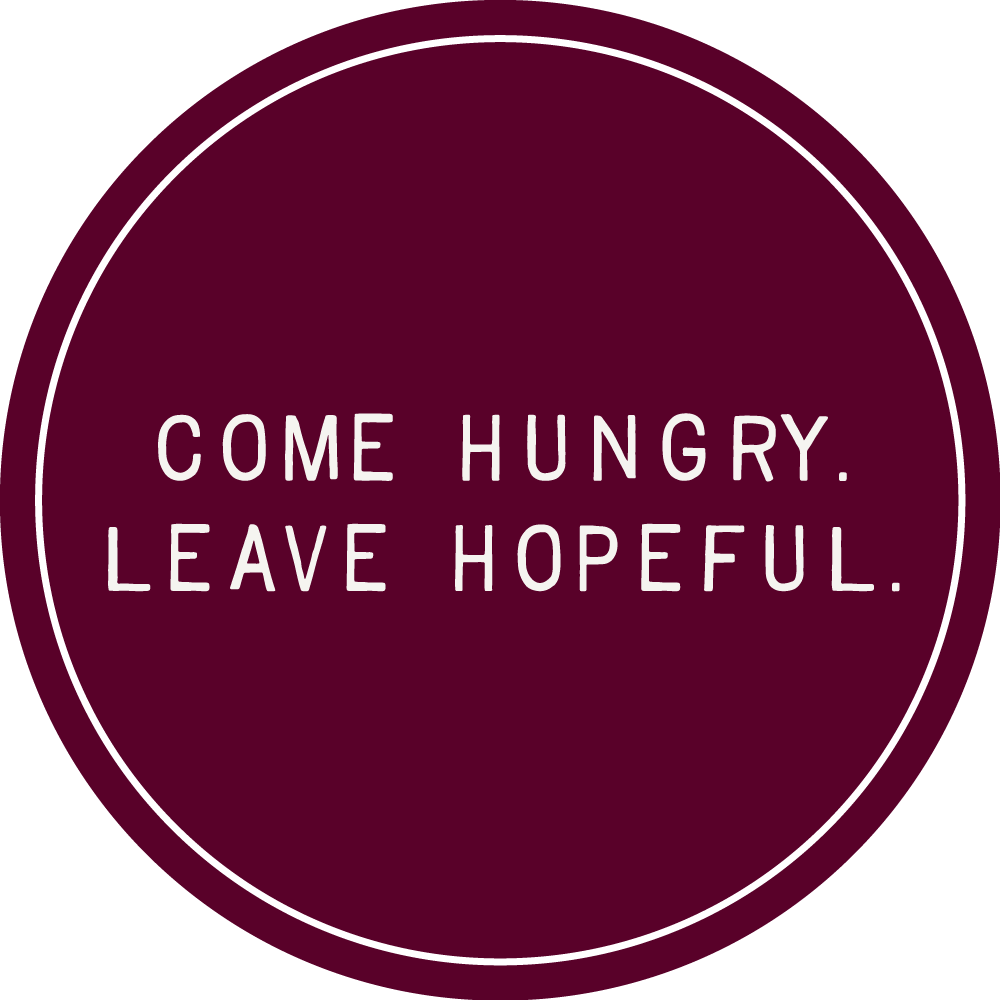 A circular sign with the quote 'Come hungry. Leave hopeful.' written in white letters on a maroon background.