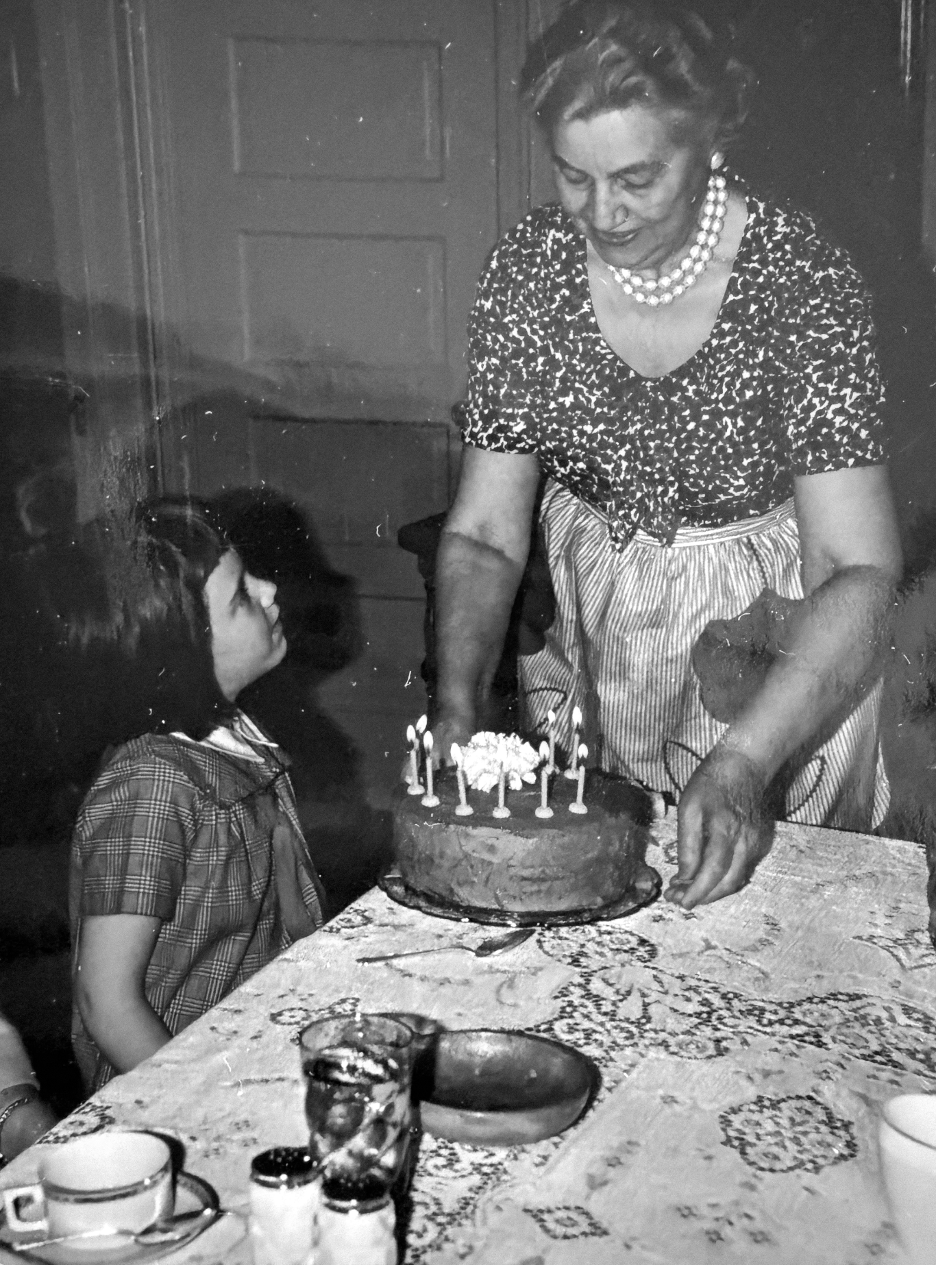 A woman is celebrating a birthday with a cake and a young girl watching; the woman is placing candles on the cake.