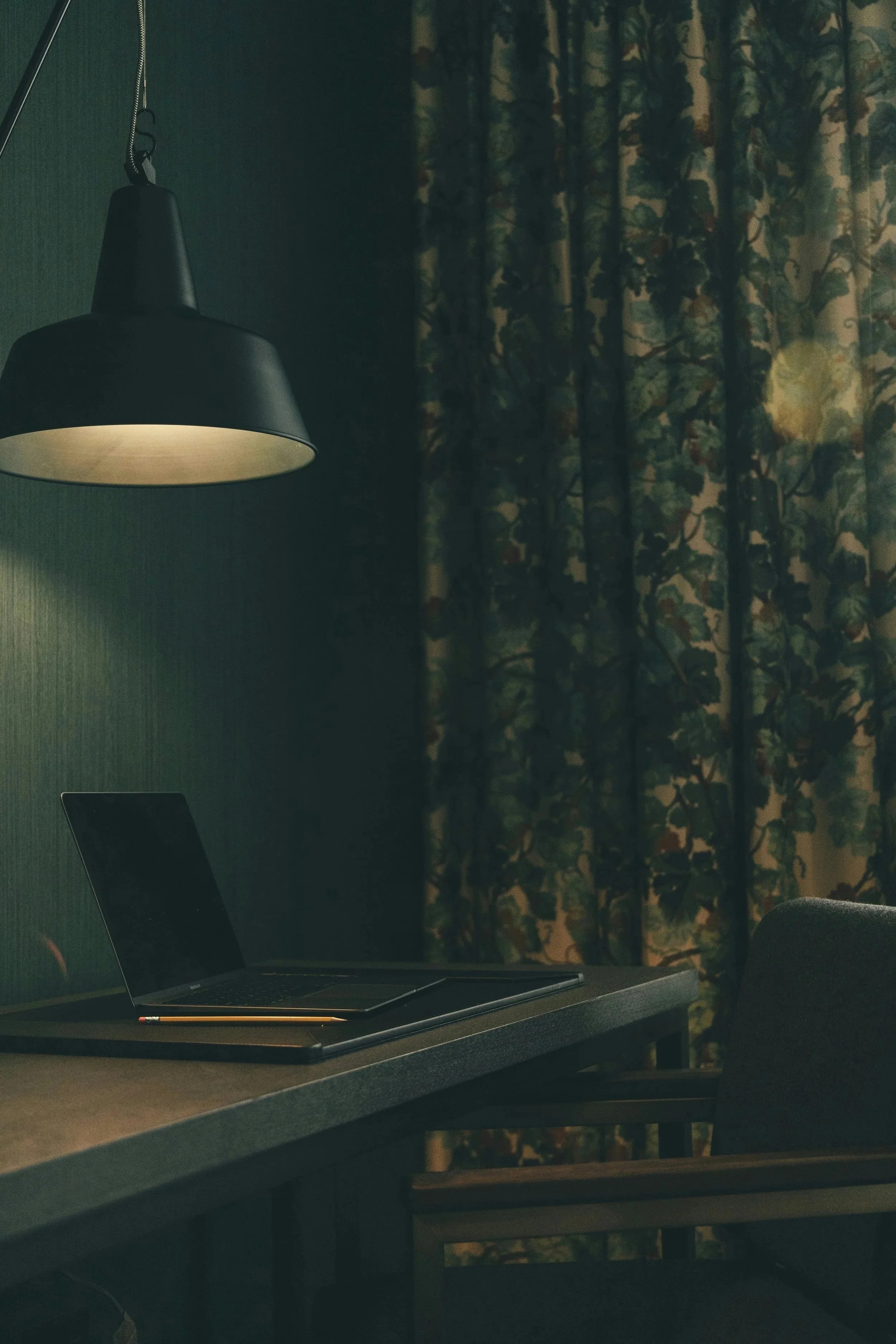 black pendant lamp glowing softly above a dark wooden desk with a closed laptop and pencil, beside a window with floral curtains.