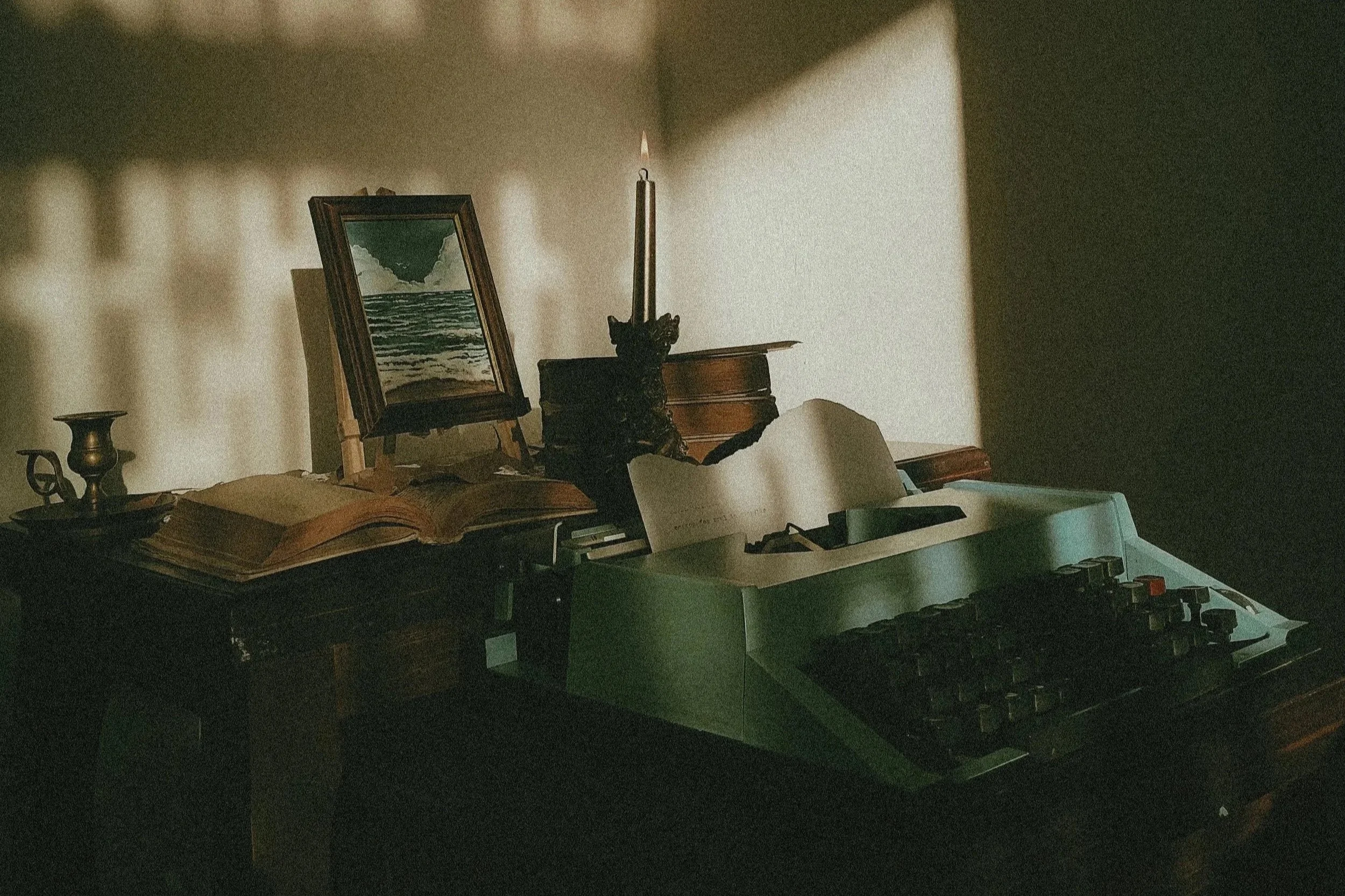 A lit taper candle in a brass holder beside an open book and folded paper, softly lit against a textured wall.