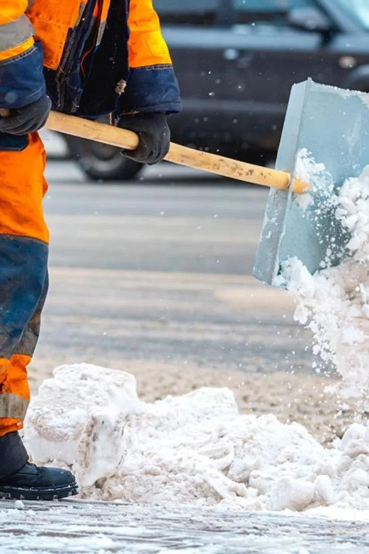 A worker in an orange jacket and black gloves shoveling snow from a street with a snow shovel.