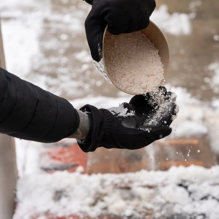 Person in black gloves pouring salt from a metal container onto snow.
