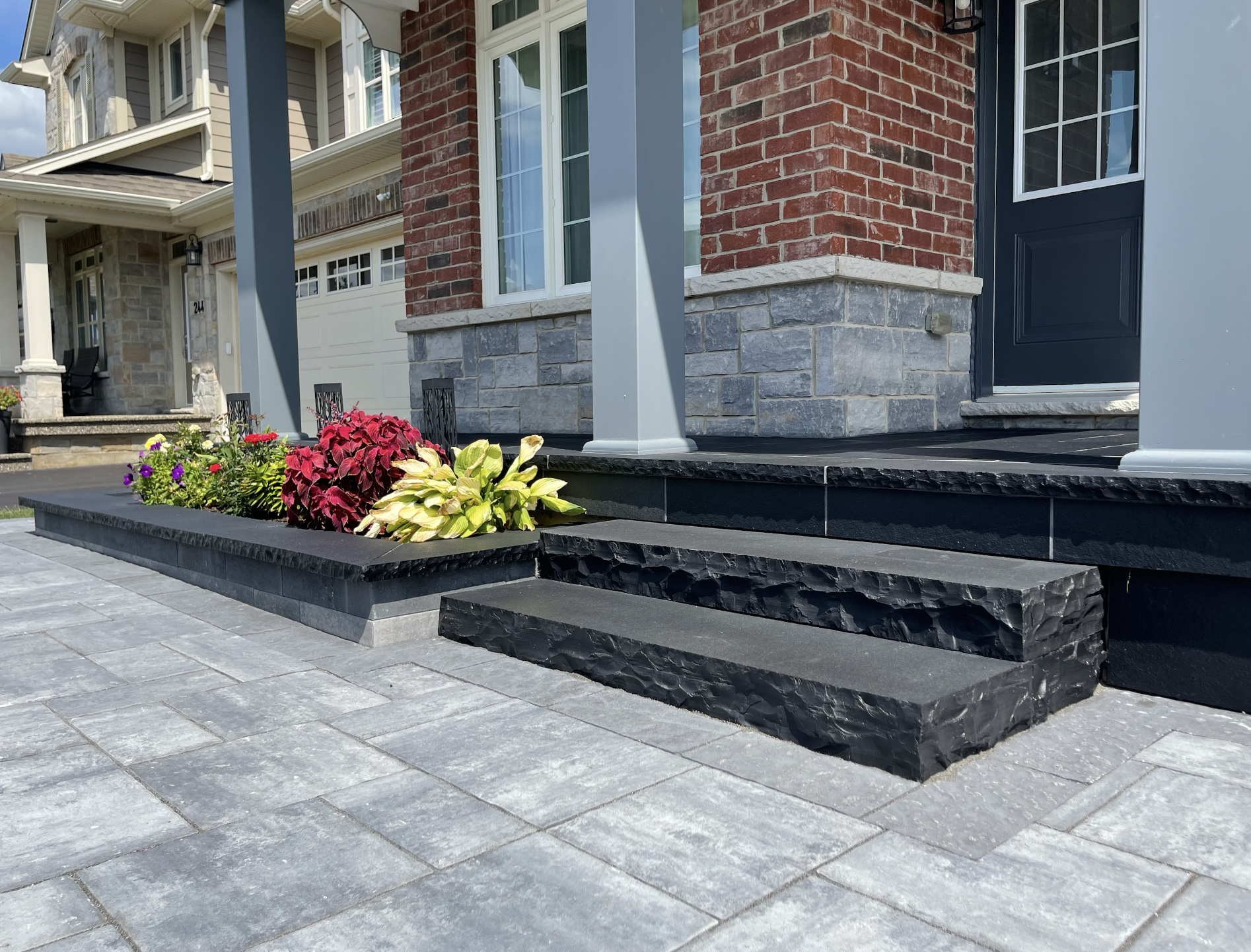 Front porch steps of a house with black stone steps, a small garden bed with colorful flowers, and a brick and stone facade.